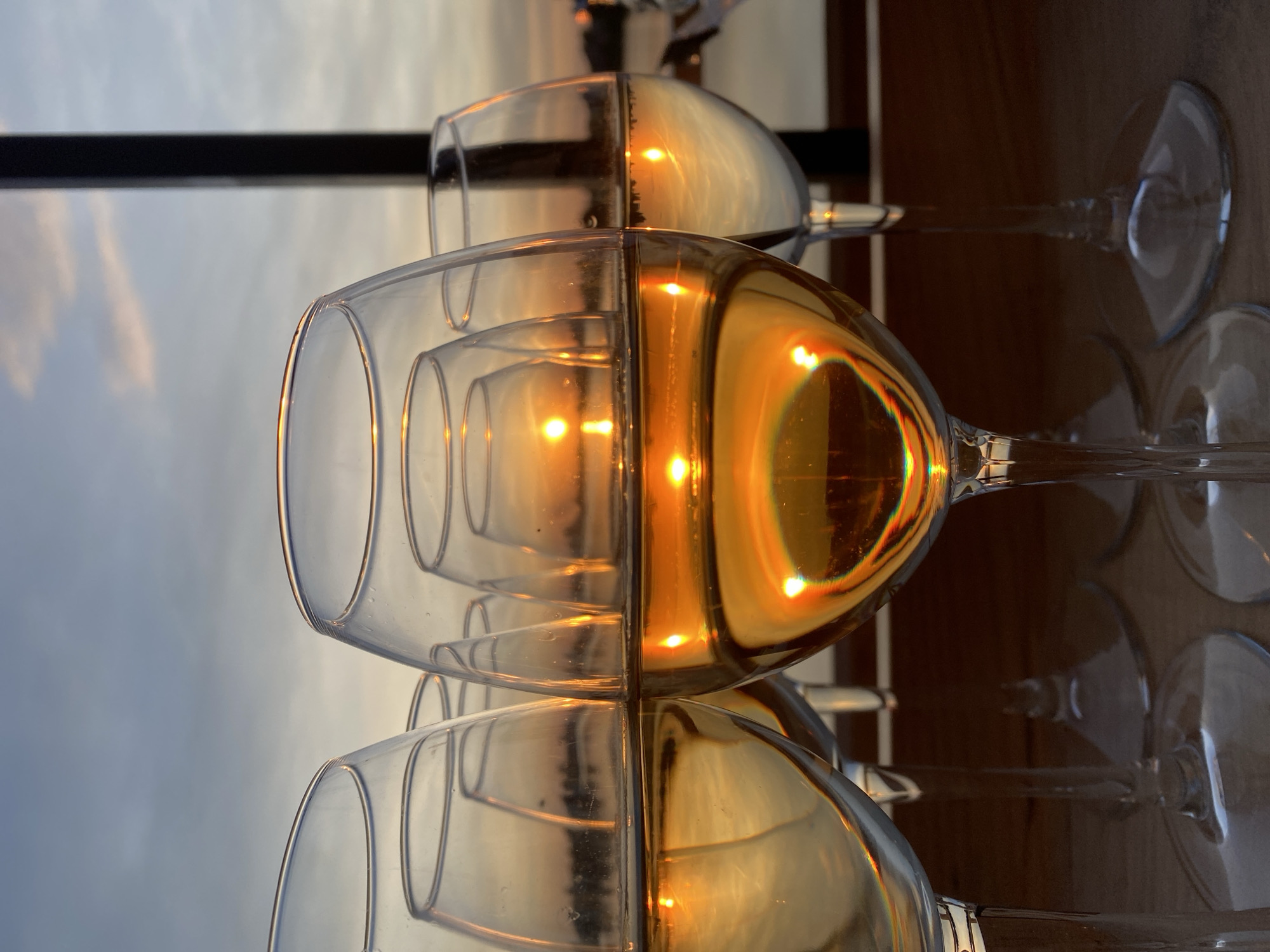 The image shows a row of wine glasses partially filled with a light-colored liquid, likely white wine, set against a backdrop of a twilight or sunset sky. The glasses are arranged horizontally, and the light from the setting sun is reflected in the wine, creating a golden hue. The glasses are clean and transparent. In the background, a dark horizontal line may represent a railing or a structural element. The setting suggests an outdoor location, possibly a restaurant or a bar with a view. The exact location within the city of Aramil, Russia, cannot be determined from the visual data.