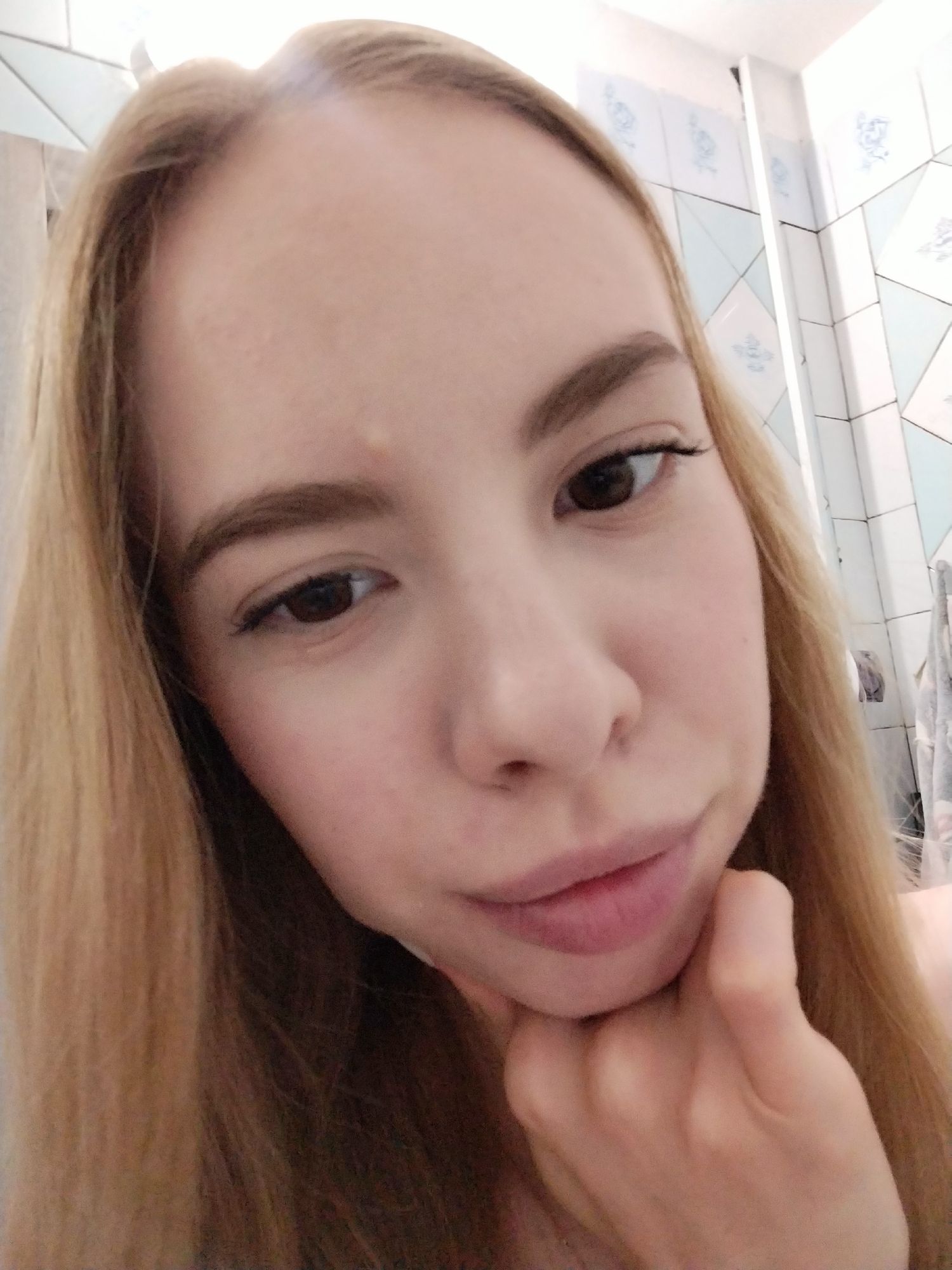 A young woman with long, blonde hair is shown in a close-up, selfie-style photograph. Her head is tilted, and her chin is resting on her hand. Her eyes are dark brown and she is looking down and to the left of the camera. She is wearing light makeup. The background appears to be a bathroom with tiled walls. The lighting is bright, and there is a slight glare on her forehead, suggesting overhead lighting. The focus is on her face, with the background slightly blurred.