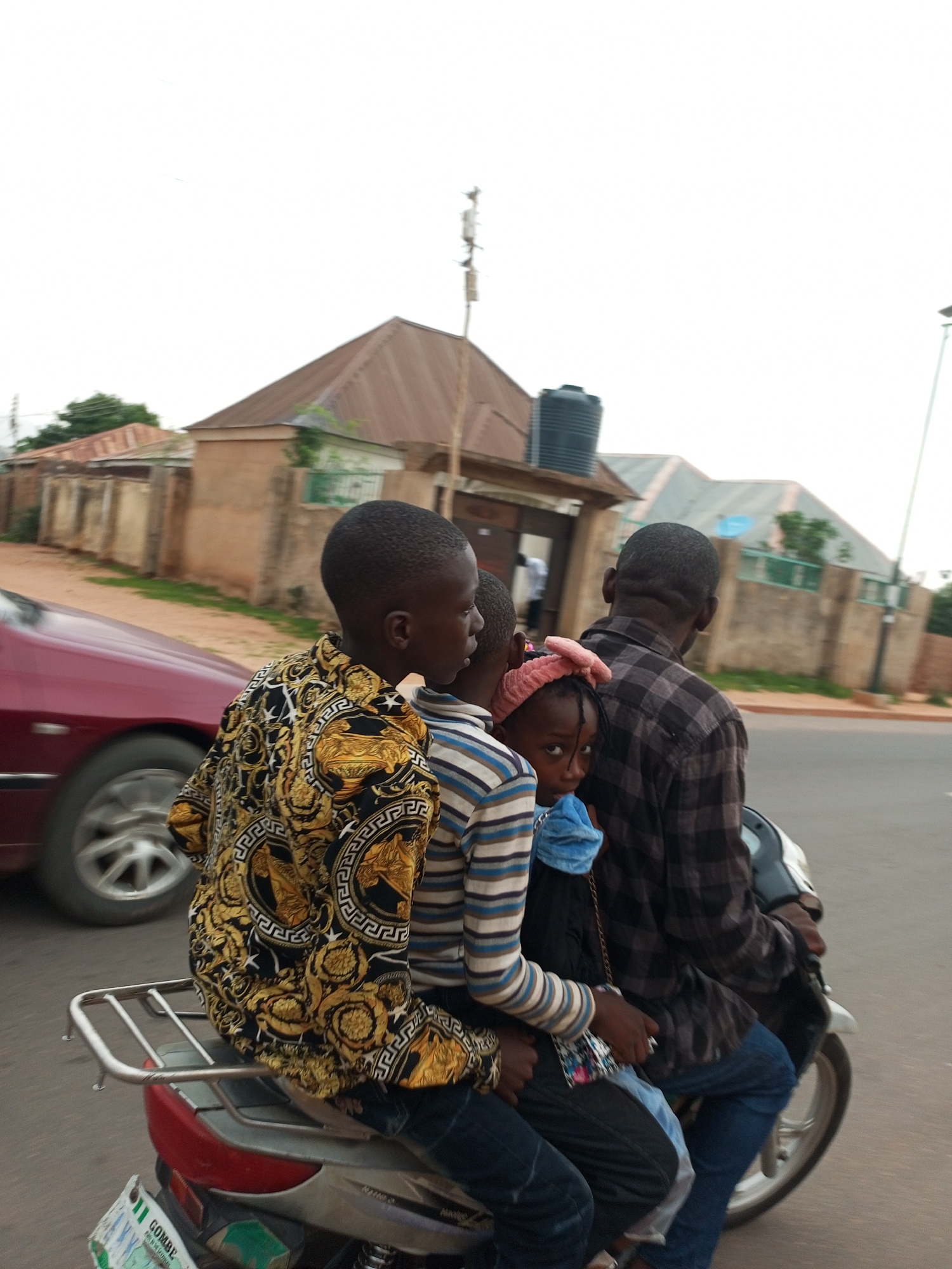 In the city of Gombe, Nigeria, four individuals are riding on a motorcycle. A man in a checkered shirt is driving, with three children seated in front of him. The child closest to the driver is wearing a striped shirt. Next to that child is a girl with a pink headband and a blue scarf. The child closest to the front is wearing a black and gold patterned shirt. The motorcycle has a metal rack on the back and a license plate that is partially visible. The background shows buildings with brown roofs and a red car is visible on the left side of the image. The sky is overcast.