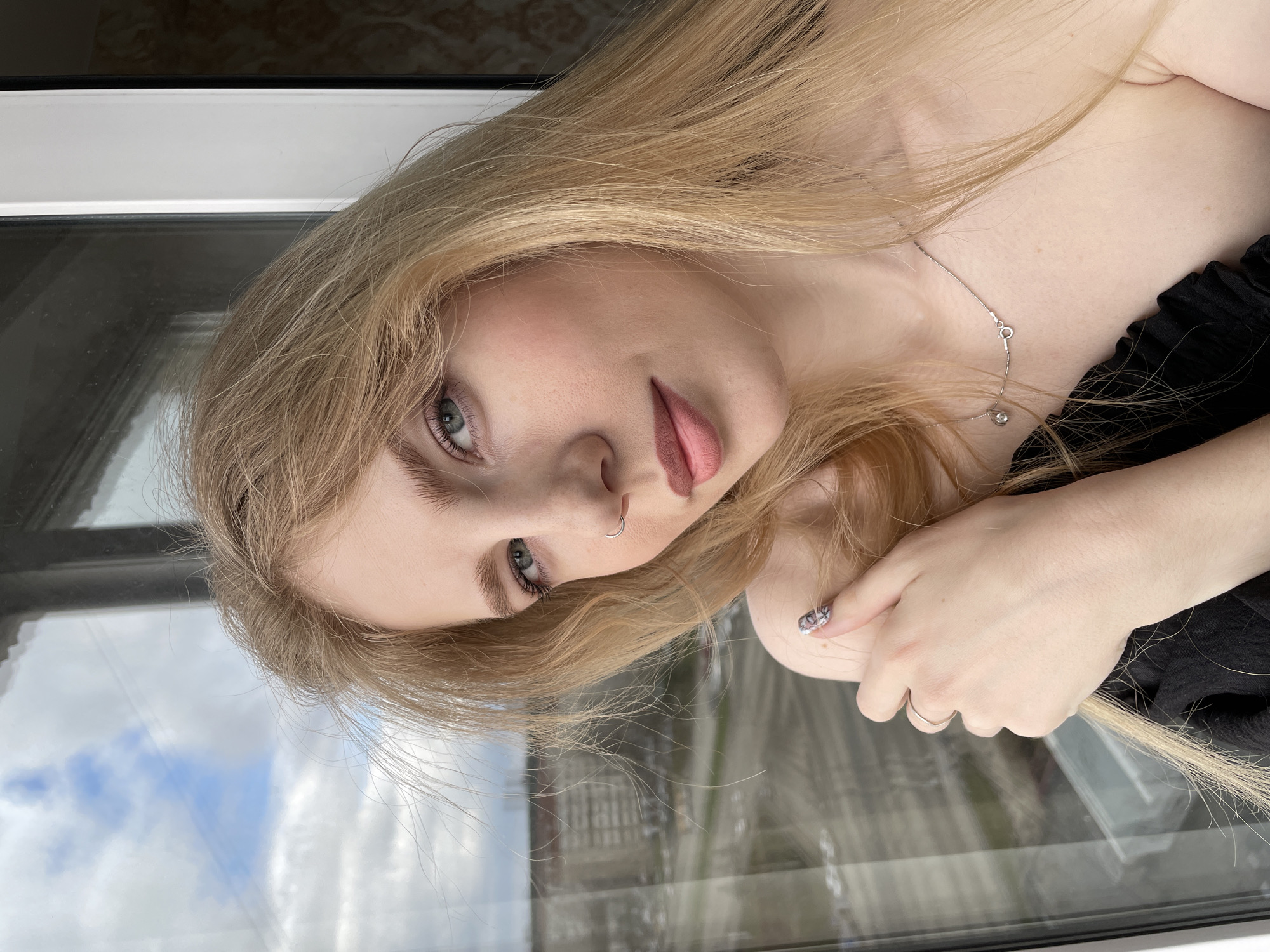 A portrait features a young woman with long, flowing blonde hair and striking light-colored eyes, possibly blue or grey-green. The image is rotated, giving the impression that she is reclining or viewed from an unconventional angle. She has fair skin, a small silver hoop nose piercing, and her lips are slightly parted in a subtle pout. A delicate silver chain with a small pendant rests on her collarbone, and a simple silver-toned ring is visible on one of her fingers. Her nails feature a distinctive marbled or abstract design. She wears a dark-colored garment, possibly a black top or dress, with ruffled details visible on the shoulder.
The woman is positioned indoors, close to a large window with a white frame. The window prominently reflects a daytime sky filled with white clouds and the outlines of distant buildings, suggesting an urban or suburban setting outside in Izhevsk, Russia. The natural light illuminates her features and hair. Her gaze is direct and composed, creating a serene yet engaging expression. There is no visible text in the image.