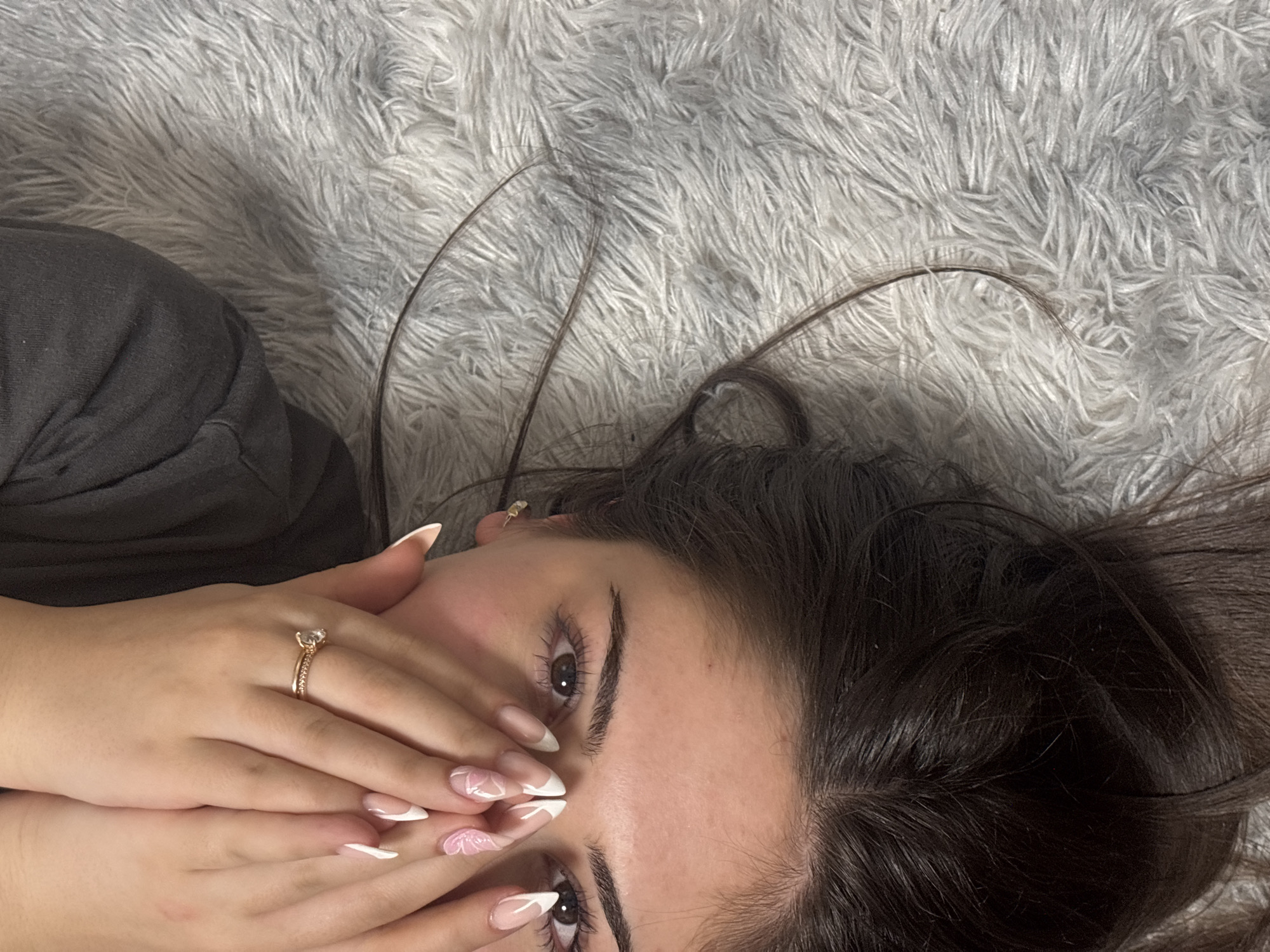 A young woman lies on a grey, fluffy surface. Her head is oriented towards the top right of the image, and her body is positioned towards the left. She has dark brown hair and brown eyes. Her hands, adorned with long, manicured nails featuring white tips and floral designs, are covering her mouth. A gold ring with a large gemstone is visible on her left ring finger. She is wearing a dark-colored garment on her left shoulder. The background consists of the grey, fluffy surface. The image does not provide any information about the city of Lehchevo, Bulgaria, or suggest it is related.
