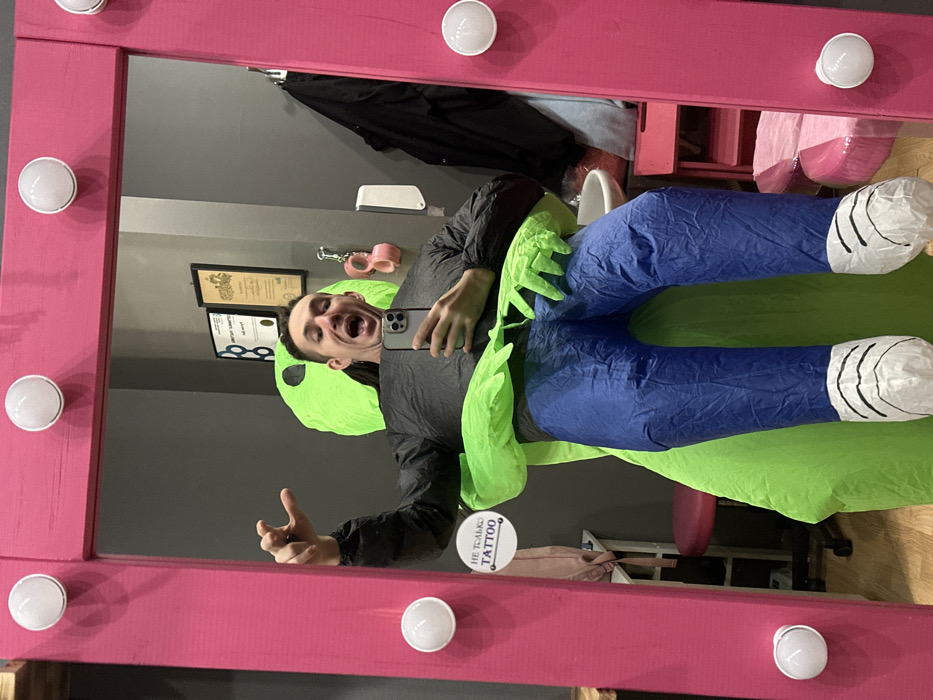 A male individual is observed taking a mirror selfie inside what appears to be a tattoo and body modification studio in Saratov, Russia.
**Scene:** The subject, a male with short dark hair and a visibly joyful, open-mouthed expression, is positioned centrally in front of a large, rectangular mirror framed in bright pink wood, illuminated by nine round, white LED bulbs. He holds a silver-colored smartphone (likely an iPhone) with his right hand, aimed at his reflection. His left arm is raised and bent, with the hand forming a loose claw shape.
**Attire:** The individual is wearing an inflatable costume. The upper portion of the costume is a dark grey or black long-sleeved garment, depicting the wearer's torso. From this garment, a bright green, presumably alien, figure extends, with large, three-fingered hands appearing to embrace the individual's waist. The lower portion of the costume consists of inflatable bright blue "jeans" terminating in white "shoes" with black stripe detailing, resembling cartoon footwear.
**Environment (Reflected in Mirror):** The background walls are painted a uniform grey. To the left, a round white sign with a pink border displays Cyrillic text: "НЕ ТОЛЬКО TATTOO" (Not Only TATTOO). Above this, a smaller, framed document is partially visible. Further to the right, two additional framed documents or certificates hang on the wall. Below these documents, a pair of pink, soft hand restraints or handcuffs hang from a hook. A white wall-mounted dispenser is located below the handcuffs, above a white ceramic basin or sink. To the immediate right of the sink, a dark jacket or garment hangs. Furniture in the reflected background includes a pink swivel stool with black wheels to the left, and a reddish-brown wooden stool or table partially visible below the sink. The overall impression is that of a professional, albeit stylized, commercial interior.