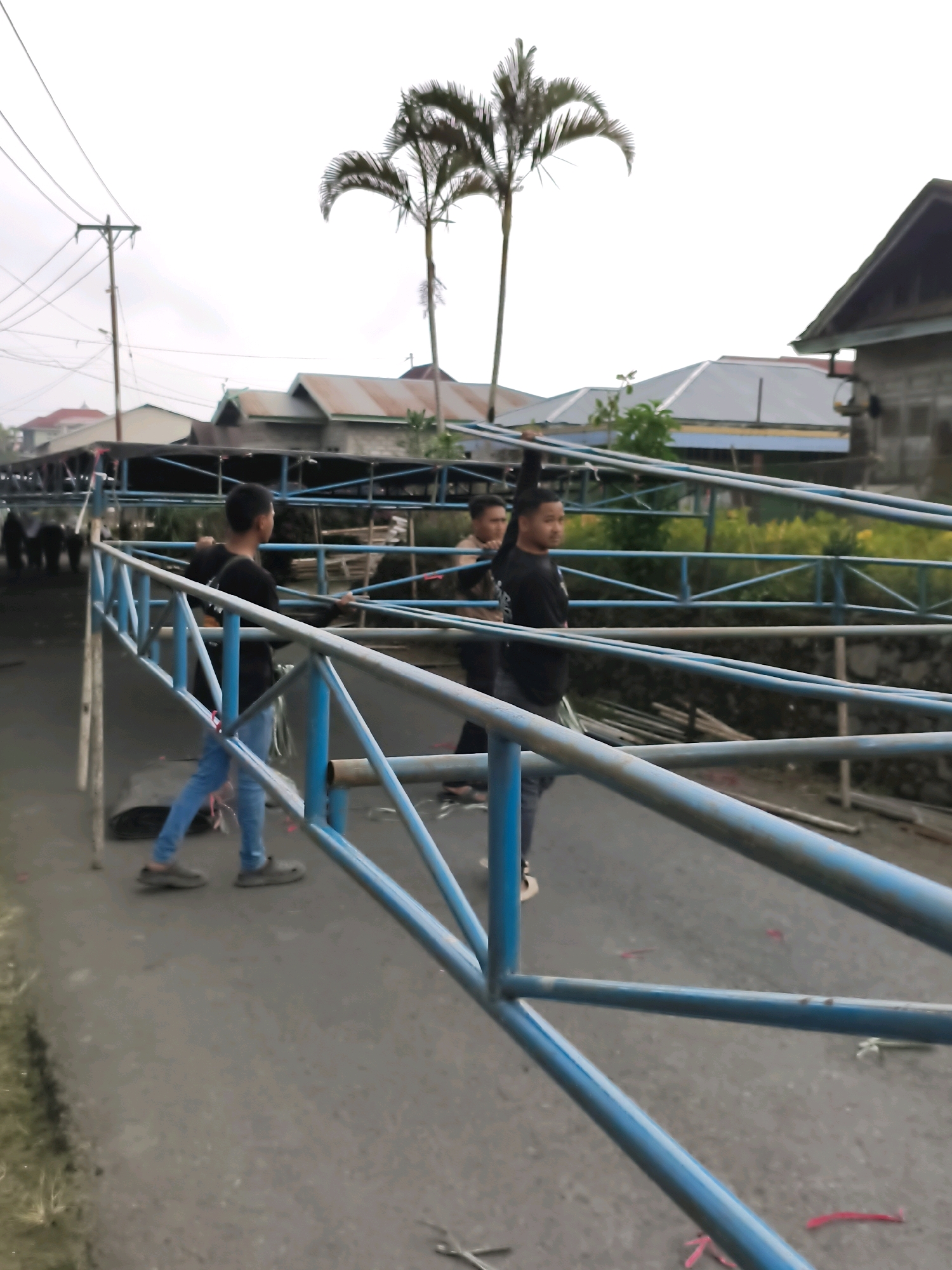 The image shows a street scene in Lindung Jaya, Indonesia, with a blue metal structure that appears to be under construction or disassembly. Three young men are visible, interacting with the structure. One man in the foreground, wearing a black t-shirt and blue jeans, is walking and looking to his right. To his left and slightly behind him, another man in a black t-shirt and blue jeans is also visible, looking towards the same direction. Further back, a third man, also in black, is seen with his back partially turned to the camera. The background shows several buildings with corrugated metal roofs and some vegetation, including two palm trees. Power lines and a utility pole are present on the left side of the frame. The ground is paved with asphalt and shows some signs of wear and debris. The lighting suggests an overcast day.