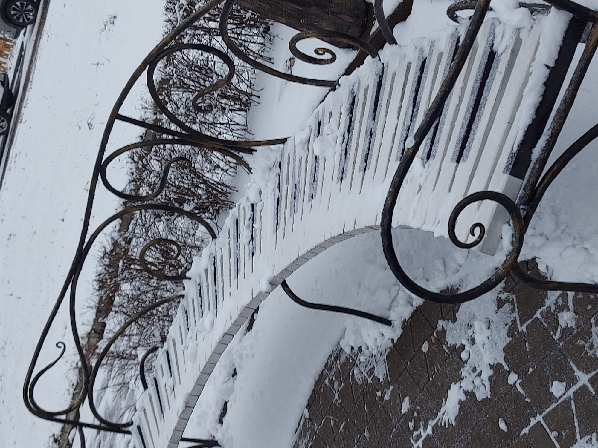 A wrought-iron bench sits in a snowy outdoor environment, likely in winter. The bench is partially covered in snow, emphasizing the cold conditions. The surrounding area also has snow covering bushes and ground, and part of a car is visible. The bench is positioned on a brick surface. The scene may be in Chaikovsky, Russia, given that location was requested in the prompt.