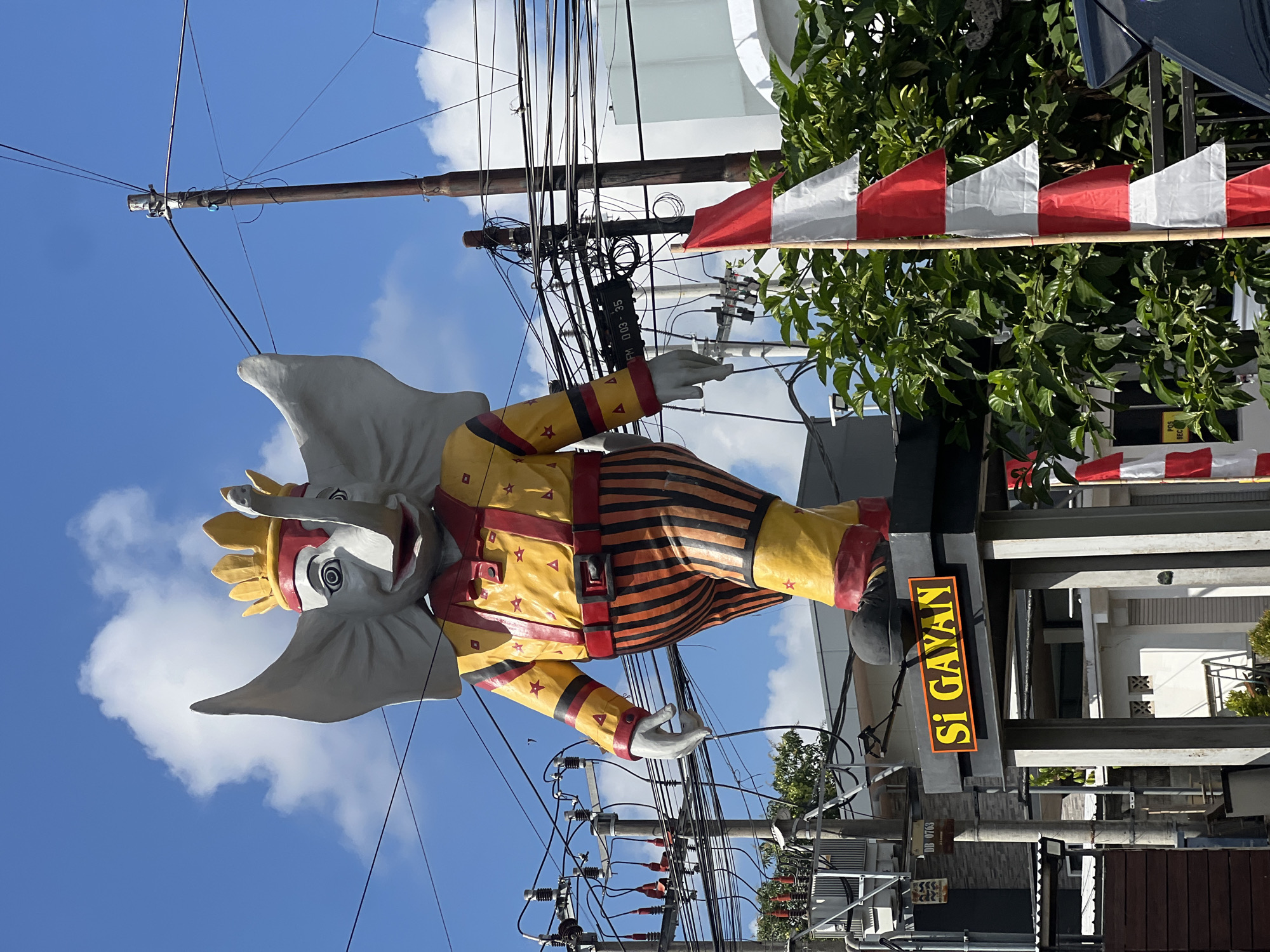 An outdoor, eye-level, medium shot captures a whimsical, oversized statue of a kingly elephant prominently displayed against a bright blue sky with scattered white clouds. The elephant, adorned in a yellow and red striped suit with red suspenders and a golden crown, stands on a grey pedestal. The pedestal features a yellow sign with the text "Si GAYAN" in brown capital letters. Below the elephant statue, electrical poles and a dense network of power lines crisscross the frame, creating a stark contrast with the vibrant colors of the statue. To the right of the statue, a bamboo pole holds a string of red and white triangular flags, reminiscent of the Indonesian flag. Lush green foliage is visible behind the flags and to the right of the statue, suggesting a tropical environment. In the lower left, a segment of an electrical transformer station is visible. The overall impression is a cheerful, yet slightly chaotic urban scene. This statue is located in Denpasar, Bali, Indonesia, and is a well-known landmark.