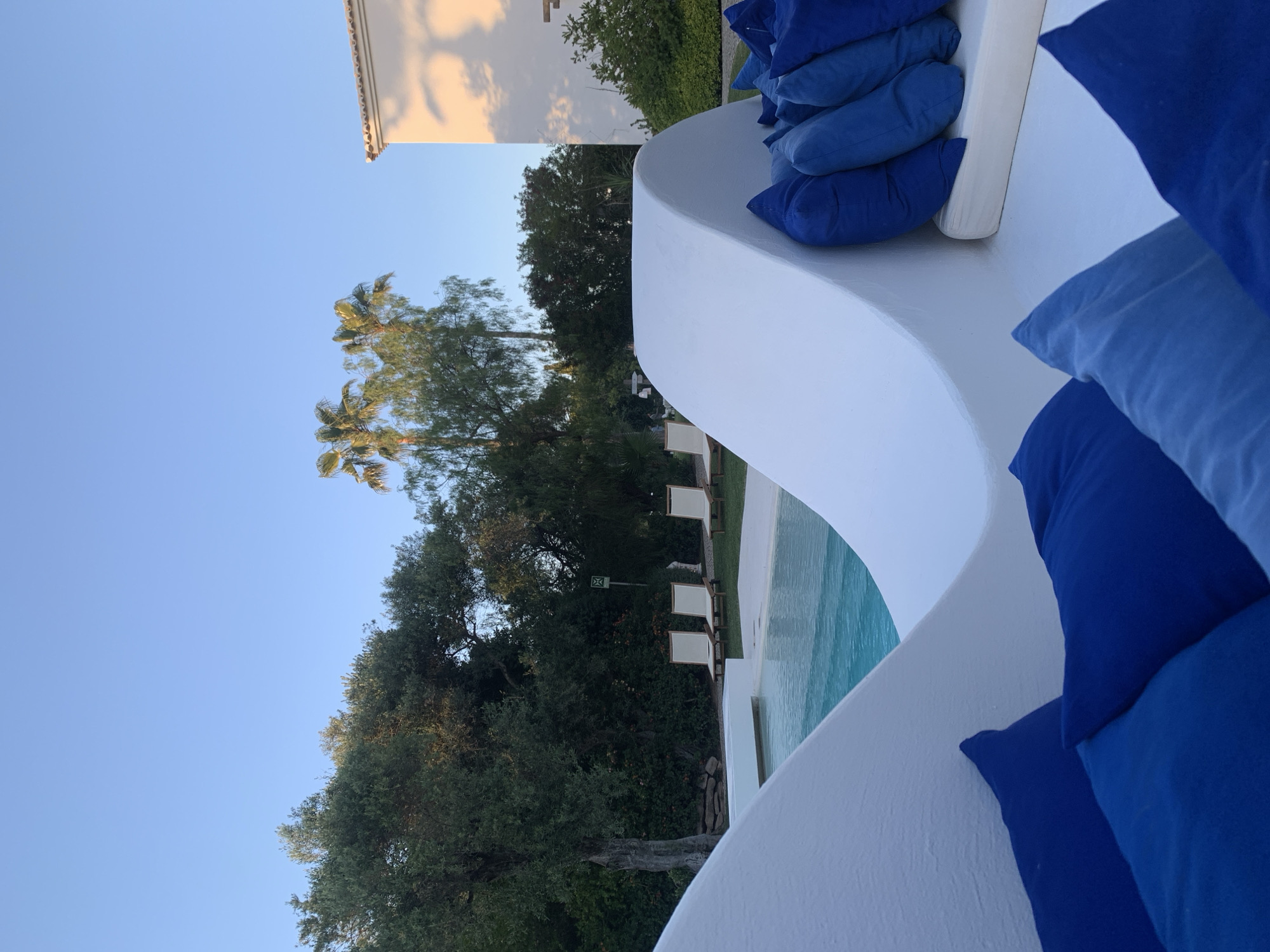 From an elevated, outdoor perspective, the image depicts a swimming pool area in Olhão, Portugal, characterized by whitewashed architecture, blue cushions, palm trees, and lounge chairs. The foreground is dominated by a curved, white wall adorned with stacks of vibrant blue cushions. Behind the wall, a swimming pool with turquoise water is visible, alongside lounge chairs neatly arranged on a manicured lawn. The background features an array of trees, including palm trees. The sky is a clear, light blue. The image suggests a serene and upscale resort or residential area within Olhão.