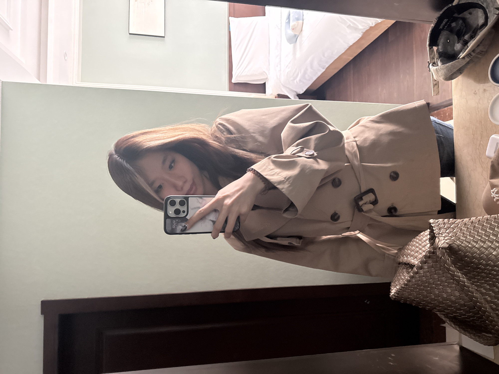 A person is taking a selfie in front of a mirror. The person is wearing a beige trench coat and holding a smartphone with a marble patterned case. Part of a woven metallic bag is visible. The reflection in the mirror shows a room with a bed, artwork on the wall, and a countertop with various items. The location is indoors, likely a hotel room or apartment. There is no confirmed information about the city of Lanzhou, China, in the image.