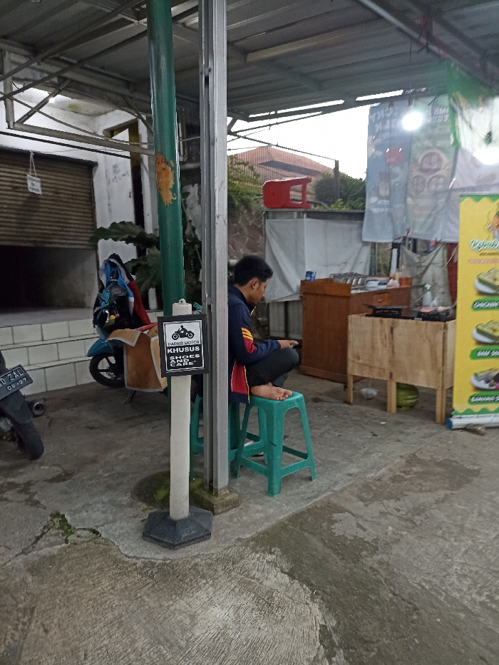 The scene takes place in Jatinangor, Indonesia, at coordinates -6.935 latitude and 107.773 longitude. It is an outdoor area with a concrete floor and a corrugated metal roof partially covering the space. A man in a black and red long-sleeved shirt is seated on a green plastic stool, facing a wooden counter with a black metal top. His feet are bare. There are two motorcycles parked in the area, one to the man's left and one to the viewer's left. The motorcycle to the left of the viewer has a license plate that reads "D-2411-ZJ." A sign with black lettering on a white background is attached to a green metal pole behind the man, and it reads "PARKIR MOTOR KHUSUS SHOES AND CARE." The sign has a picture of a motorcycle at the top. The counter is partially covered with a white sheet. On top of the counter, there is a black metal grid and several other unidentified objects. A green metal pole is located behind the man, and a white pole is located to the man's left. A small pile of dirt is located on the ground behind the man, and the ground around the man is dry and cracked.