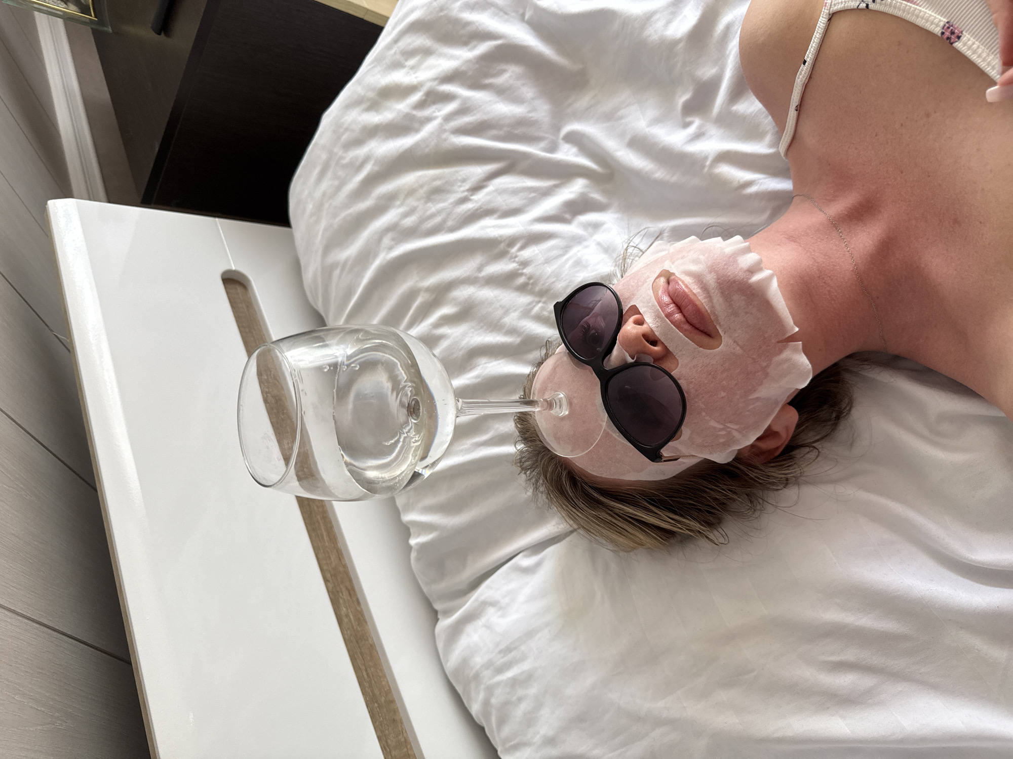 The image displays a woman lying on her back on a white bed, captured from an overhead perspective. She is engaged in a self-care routine, wearing a pinkish sheet mask on her face and black sunglasses over the mask. Humorously, a clear wine glass filled with water is balanced on the bridge of her nose, resting on the sunglasses. Her lips are visible through the mask's opening.
The woman has light brown hair and is wearing a light-colored, patterned strap top or bra. A delicate silver chain necklace adorns her neck. The white bedding is slightly rumpled, indicating a comfortable and relaxed environment. In the background, a white headboard with a light wooden accent is visible on the left side of the frame, and a dark wooden bedside table is partially seen at the top. The scene takes place indoors, likely a bedroom, creating an atmosphere of leisure and lightheartedness. There is no visible text. The location context is Chaikovsky, Russia.