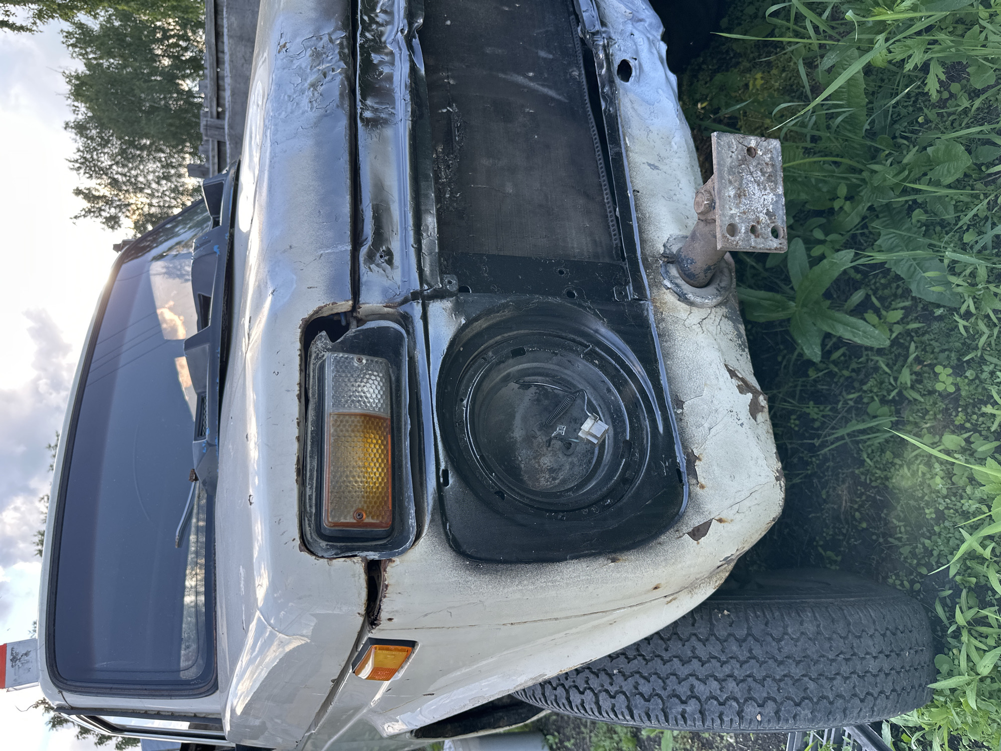 The image shows the front, partially damaged section of a white off-road vehicle, possibly a Lada Niva, parked outdoors on grassy ground. The vehicle's paint is weathered and peeling, with visible rust spots. The front grille is missing, exposing the radiator, which appears damaged. The right headlight housing is intact and contains an amber turn signal and a clear parking light. The left headlight housing is missing, revealing the empty socket for the bulb. A tow hitch is mounted on the lower front bumper. The sky in the background is partly cloudy, with the sun setting or rising, casting a warm light. Trees and a wooden fence are visible behind the vehicle. The scene appears to be in a rural or unkempt area, not within a city.