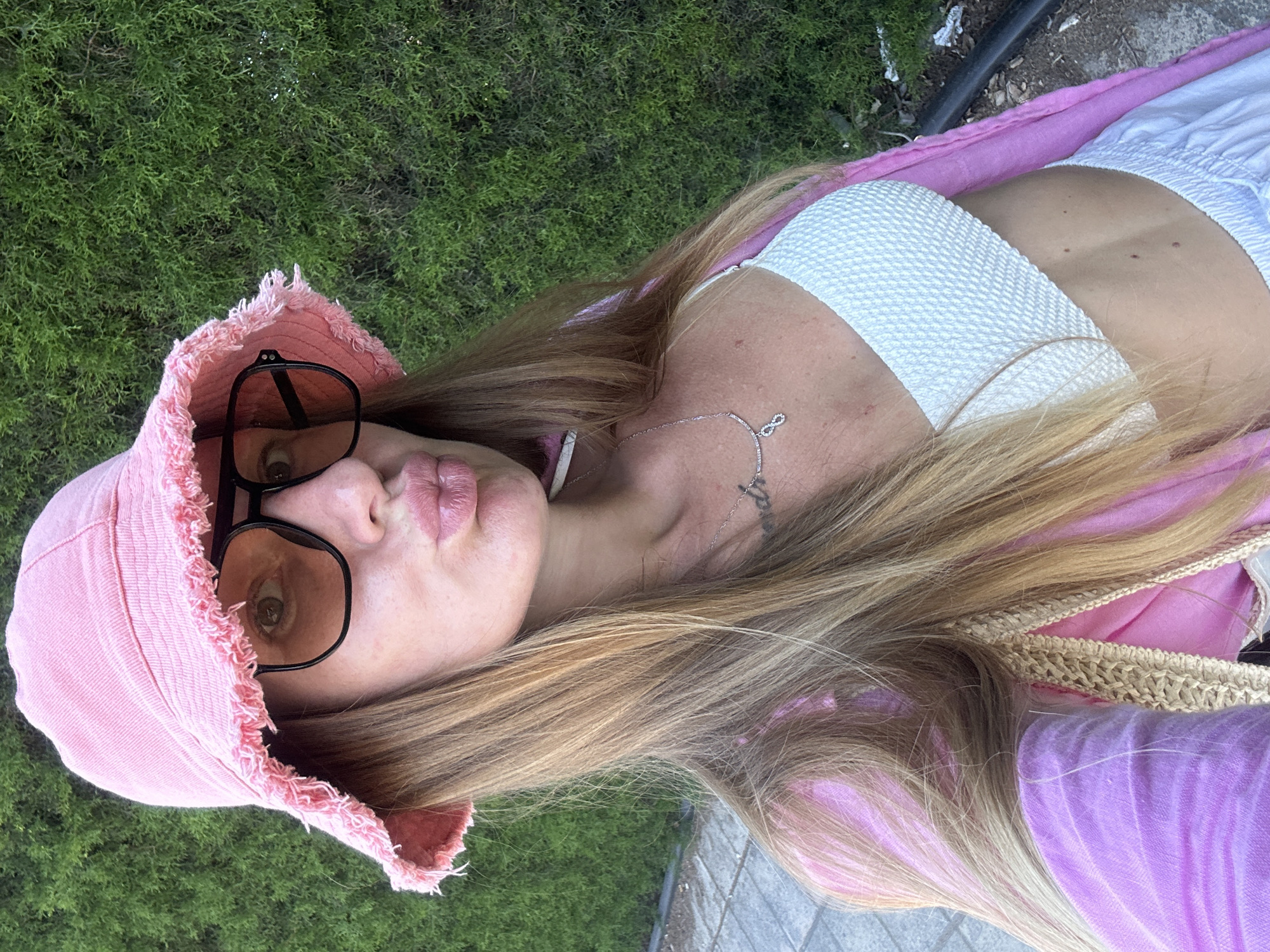 A woman wearing a pink bucket hat and sunglasses with orange lenses is taking a selfie outdoors. She has on a white textured bikini top and a pink open-front garment. Her long blonde hair is visible, framing her face as she pouts her lips. A silver necklace with an infinity symbol pendant is visible on her neck. Behind her is a lush green hedge. The ground in front of her appears to be a paved walkway. The scene is set in the city of Peyia, Cyprus.