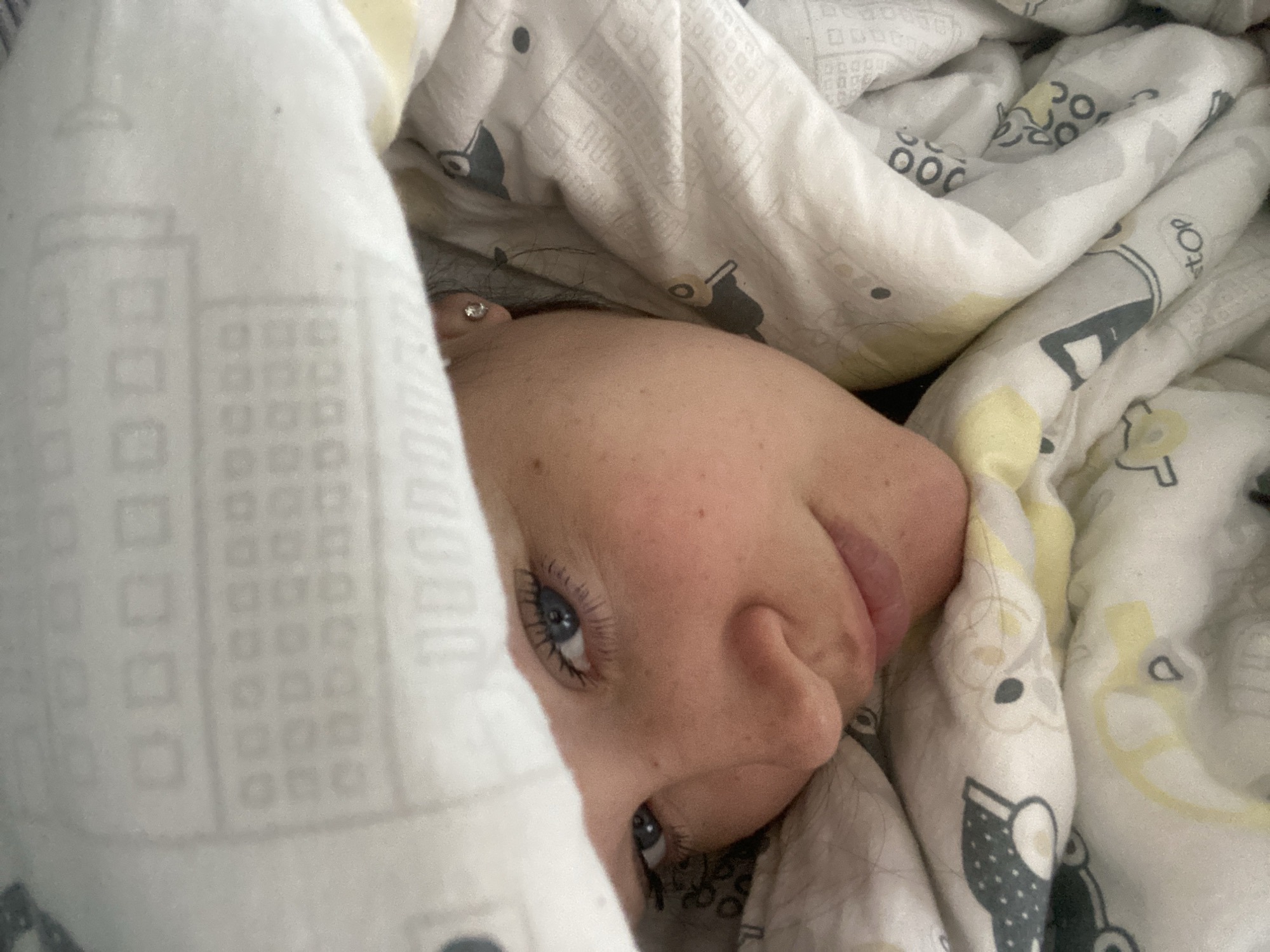 An overhead close-up shot captures a person partially hidden under white and yellow patterned bedding, appearing to be in a bed. The person's face is seen from the nose up, turned slightly to the right. One bright blue eye is open, with long dark eyelashes visible. A small portion of the other eye can also be seen. The skin has a scattering of freckles. On the visible earlobe, a small, sparkly stud earring is present. Dark hair is visible near the ear.
The bedding features a repeating pattern of stylized buildings or squares, and abstract shapes in grey and yellow. In some sections, text is discernible, including "STOP" and "JOE". The overall impression is one of coziness, possibly a morning or late-night scene. The person's expression seems calm and perhaps a little sleepy or curious, peeking out from under the covers. No other specific location cues for Sochi, Russia, are visible within the image itself.