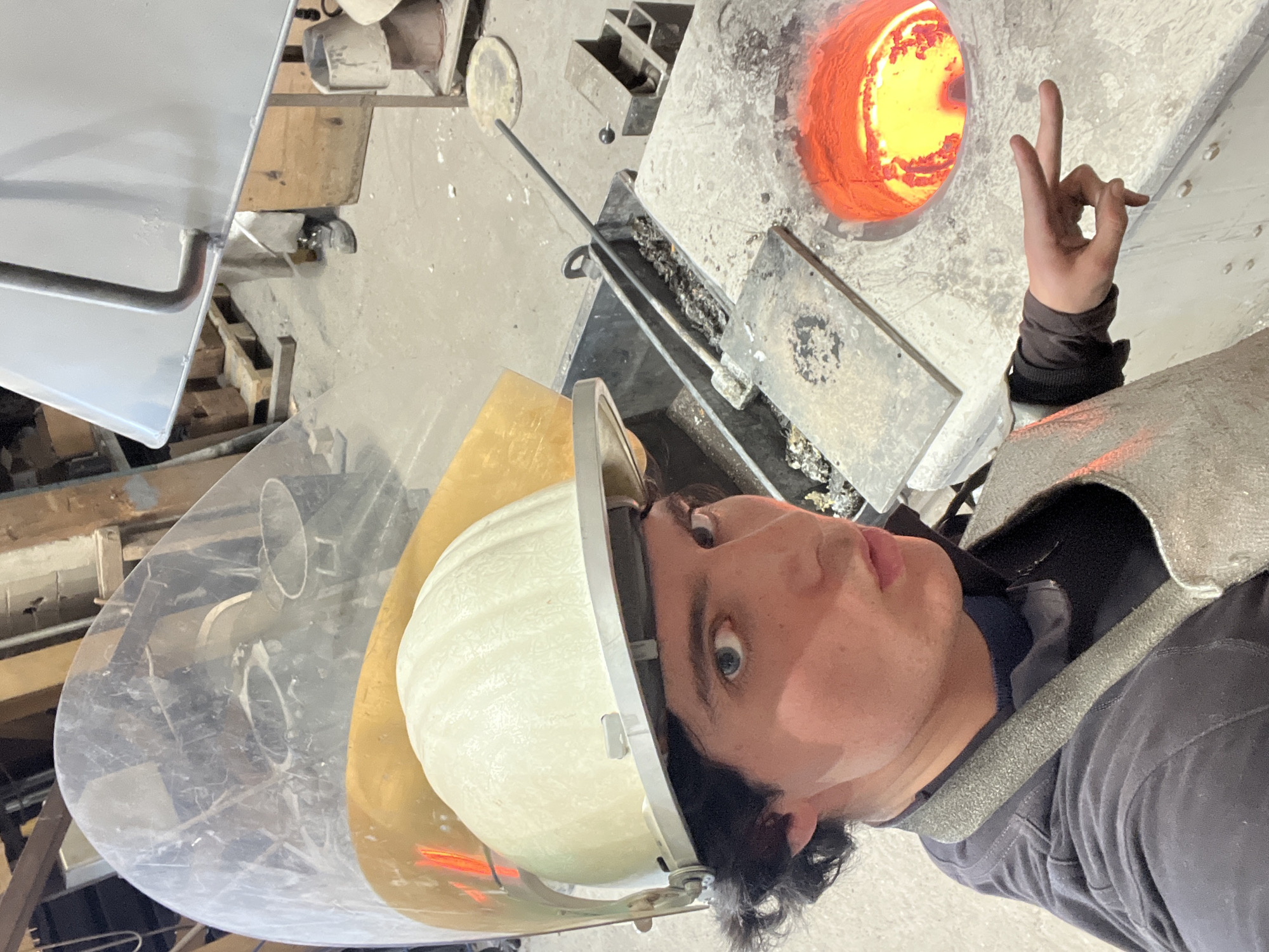 A man wearing a protective helmet with a transparent visor and a heat-resistant vest stands near an open furnace. The furnace appears to be industrial, containing a glowing orange-yellow light within. The man is making a gesture with his hand, holding up two fingers. Various tools and metallic objects are scattered around the furnace. The location cannot be verified as Saint-Rémy-lès-Chevreuse, France.