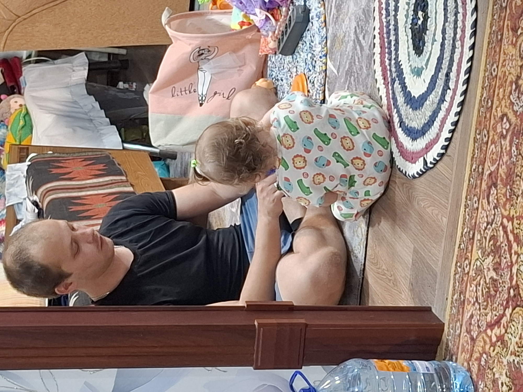 A man is sitting with a baby. The man is fair-skinned, balding, and wearing a black t-shirt. The baby is fair-skinned, wearing a onesie with cartoon animals on it, and has a small ponytail. They are sitting near a braided rug, a laundry basket, and a bottle of water. The location may be Nizhnyaya Gar', Russia, but this is unconfirmed from the image.