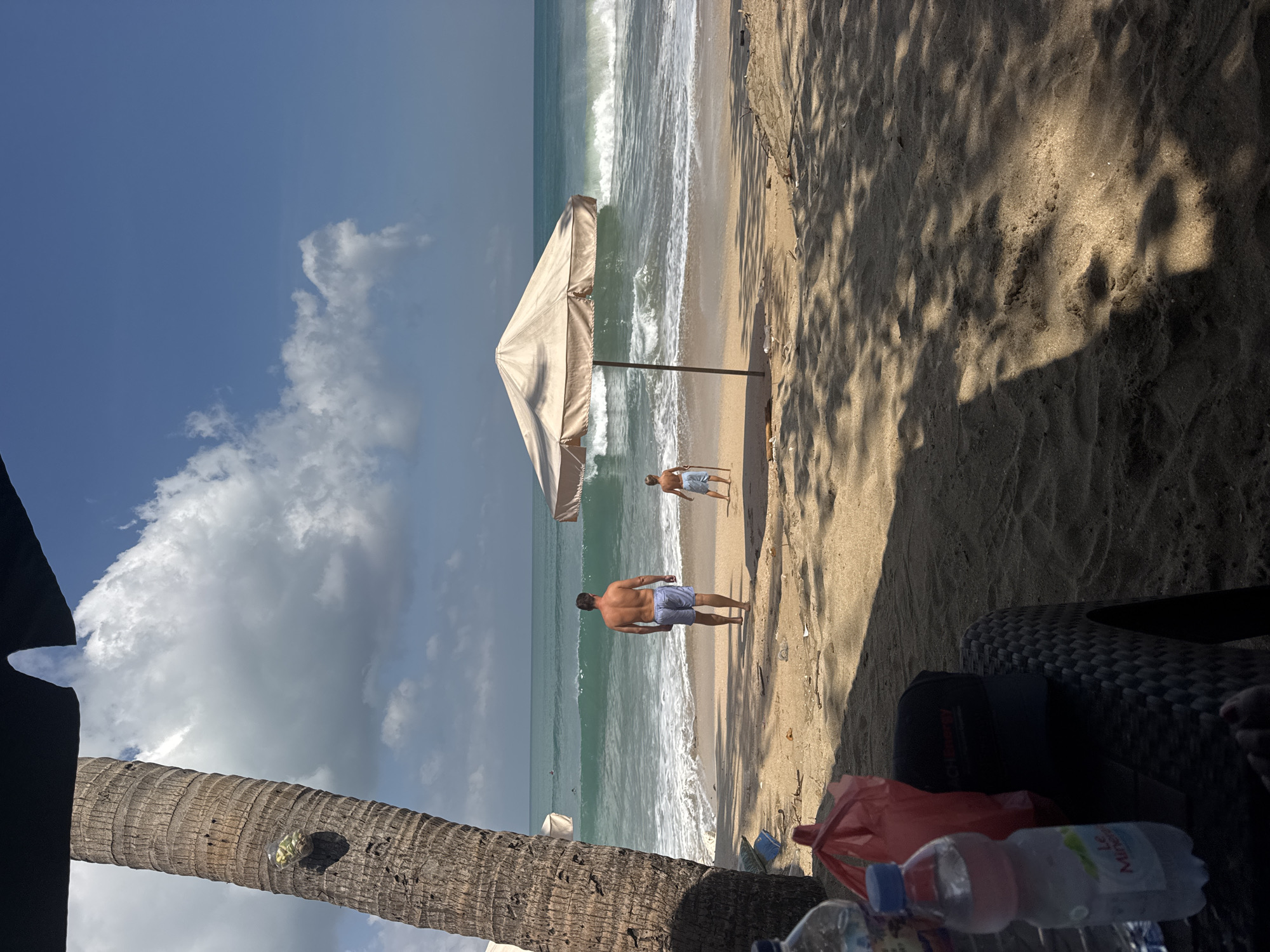 The image depicts a tropical beach scene in Kuta, Indonesia, on a sunny day. In the foreground, a woven table with items like a bottle of water, a red bag, and a black cap is partially visible. A large palm tree trunk is on the left, casting a shadow. In the middle ground, two individuals, an adult male and a younger male, are walking towards the ocean. Both are wearing swim trunks. The adult male is facing away from the camera, and the younger male is holding a stick. A large beach umbrella stands on the sand between them and the water. The ocean features moderate waves breaking near the shore, with a few surfers visible in the distance. The sky is blue with scattered white clouds. The overall environment is sandy with some scattered debris on the beach.