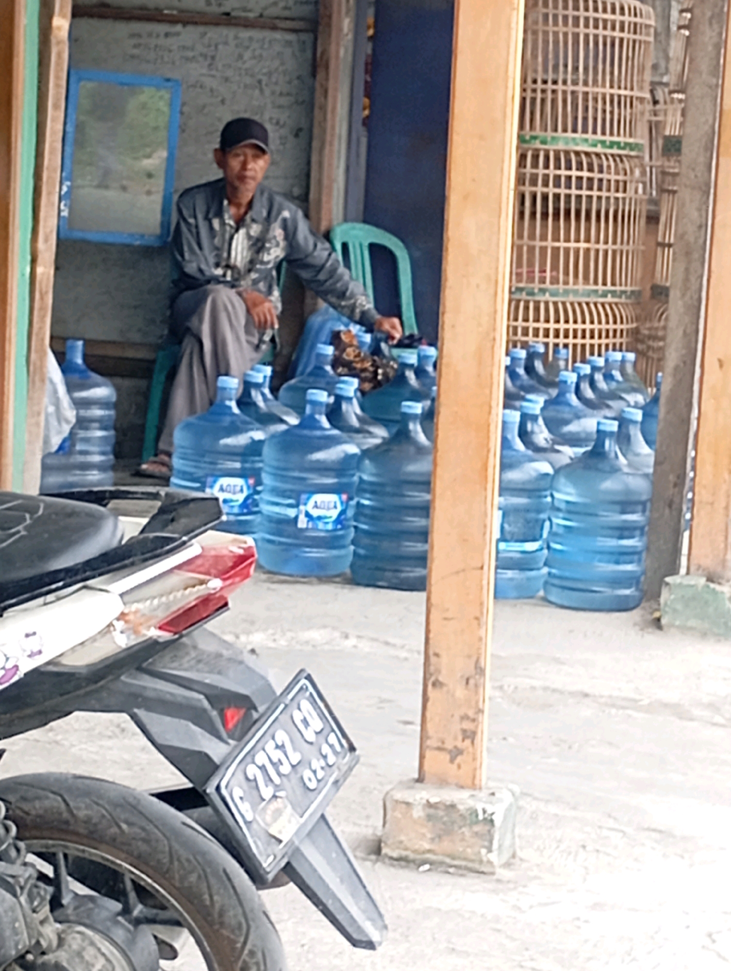 The image captures a street scene in Babelan, Indonesia, featuring a man sitting on a chair in front of a stall. He is wearing a patterned shirt, grey pants, and a black baseball cap. In the foreground, a motorcycle is parked with its rear wheel and license plate visible, which reads "G 2752 CO" and "02-27". Behind the man, numerous large blue water jugs are stacked. To the right of the jugs, several birdcages are stacked vertically. The stall appears to be an open-air establishment, with wooden posts and a concrete floor. A mirror with a blue frame hangs on the wall behind the man. The overall impression is of a typical roadside vendor setting.