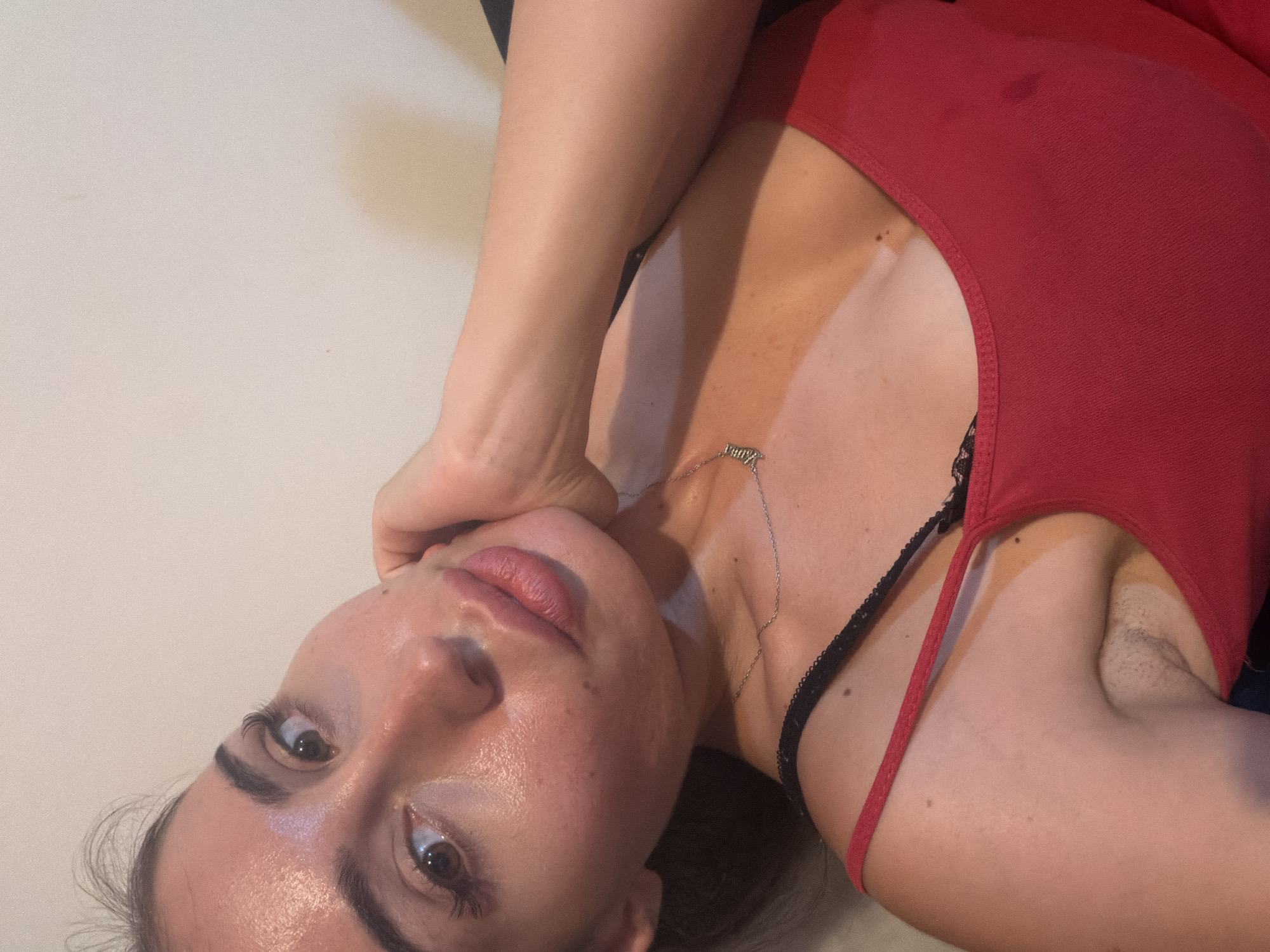 The media file shows a close-up, rotated, and slightly blurry shot of a person, likely female, wearing a red tank top and a black lace bra. The person is lying down on a light-colored surface, possibly a bed or a yoga mat. Their face is turned towards the camera, with one hand gently supporting their chin and cheek. Their eyes are open and looking directly at the viewer. They have full lips and dark eyebrows. A delicate silver necklace with a small pendant is visible on their collarbone. The scene appears to be indoors, with neutral lighting that casts subtle shadows. There is no visible text in the image. The cropped images provide closer views of the person's face, neck, shoulder, and upper torso, highlighting the textures of their skin and clothing. The setting is not definitively identifiable beyond it being an indoor space.