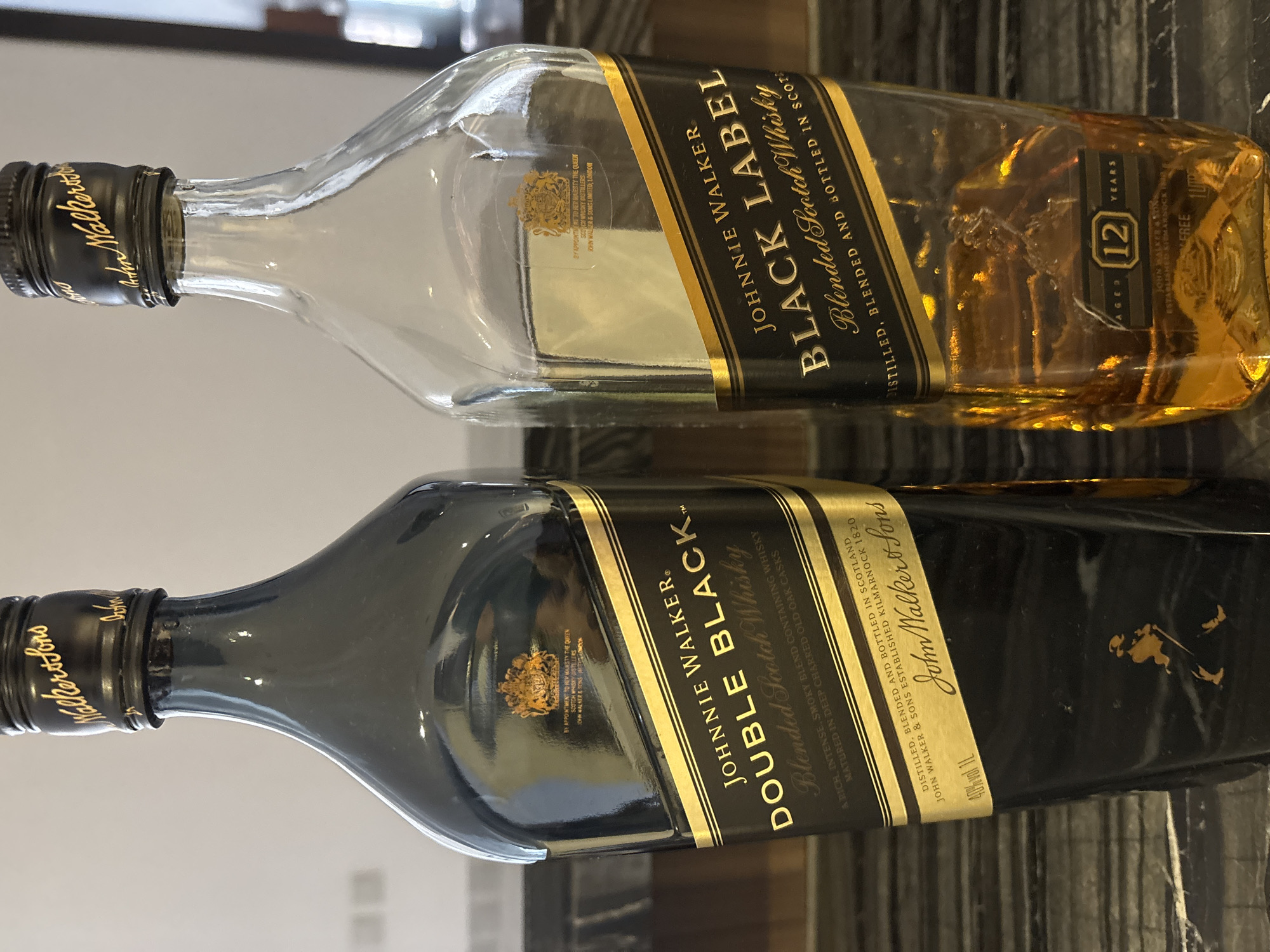 Two bottles of Johnnie Walker whisky are placed side-by-side on a dark, textured surface, likely a countertop or table. The bottle on the left is a full, opaque black bottle with a prominent black and gold label that reads "JOHNNIE WALKER DOUBLE BLACK Blended Scotch Whisky". The bottle on the right is a clear glass bottle, partially empty, with amber-colored whisky visible. Its label also reads "JOHNNIE WALKER BLACK LABEL Blended Scotch Whisky" and features a smaller label at the bottom indicating "AGED 12 YEARS". The cap of both bottles bears the "John Walker & Sons" script. The background is a neutral, lightly colored wall with some indistinct elements, suggesting an indoor setting. There is no indication of the city of Dubai, United Arab Emirates, or any specific location within it.