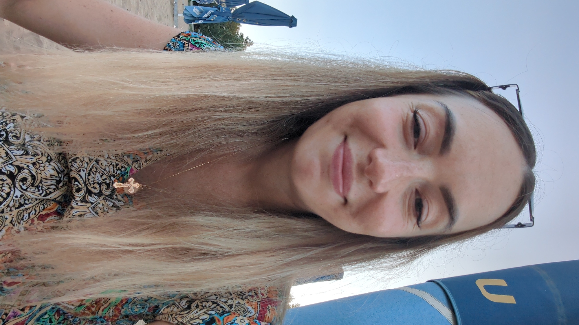 The image shows a medium shot of a young woman with long, blonde hair, wearing a colorful, patterned dress and a gold cross necklace. She is wearing glasses pushed up on her head. She is outdoors, with a clear blue sky in the background. To her right, a large blue cylindrical object with a yellow "U" is visible. In the background to her left, blue beach umbrellas are folded. The setting appears to be a beach or an outdoor recreational area, potentially near the Nistru River in Vadul lui Voda, Moldova, given the presence of beach umbrellas and the natural lighting suggesting daytime. The woman is looking directly at the camera with a slight smile.