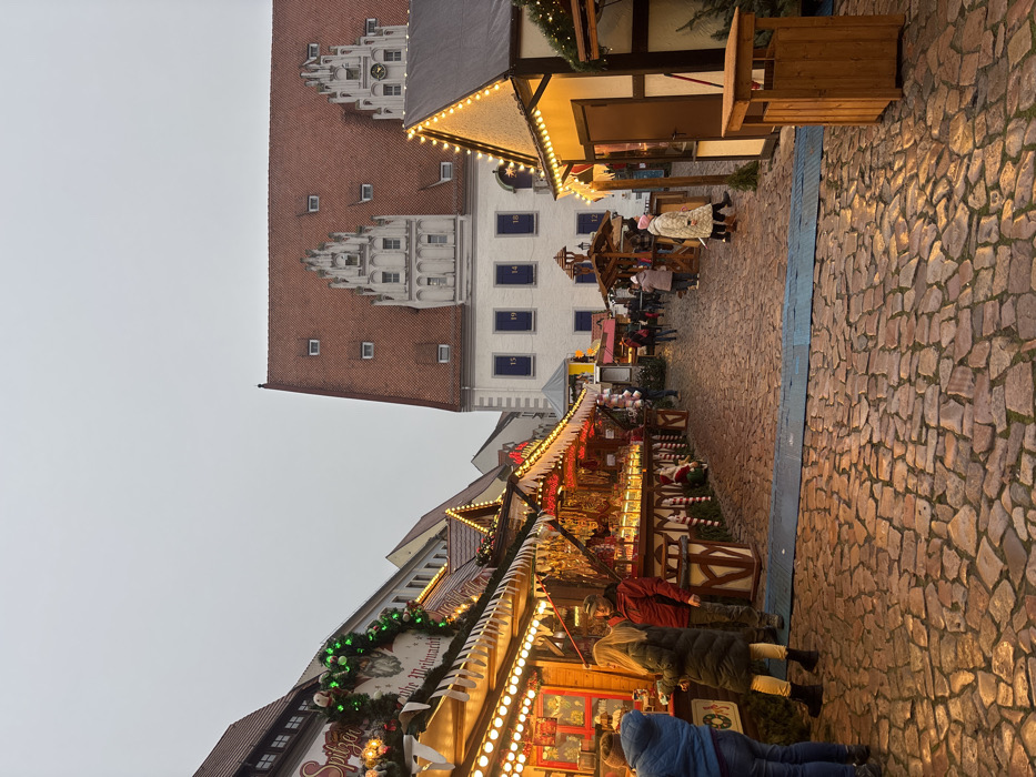 The image depicts an outdoor Christmas market scene in Meissen, Germany, specifically located in the market square in front of the Albrechtsburg. The sky is overcast and dull, suggesting late afternoon or early evening, with numerous artificial lights illuminated on market stalls.
In the foreground, the ground is paved with irregular, wet cobblestones, reflecting the warm glow from the market lights. A blue strip of material spans the width of the foreground. To the left, a series of decorated market stalls line the cobblestone path. These stalls feature white and red striped awnings, adorned with green garlands, string lights, and Christmas decorations. One stall sign partially displays "Spitzen..." and "Frohe Weihnacht." Goods, including items resembling dried food or crafts, are displayed under the illuminated awnings. Three individuals are visible at the leftmost stall: one person in a blue jacket with their back to the viewer, a woman with long blonde hair in a dark green coat, and a man in a red jacket and black beanie hat, all appearing to browse.
Further down the market path, additional stalls are visible, some with wooden frameworks and string lights. On the right side of the image, another market stall is present, with string lights and a dark wooden door. A wooden trash receptacle stands beside it.
In the background, a large, historic building with a prominent red tiled roof dominates the scene. The roof features numerous rectangular dormer windows and two ornate white gables. The building's white facade has multiple dark-framed windows, some of which bear numbers (e.g., 15, 19, 14, 18) in a blue font. This building is identified as Albrechtsburg Meissen. Beyond the immediate market area, several indistinct figures are visible walking through the cobblestone square.