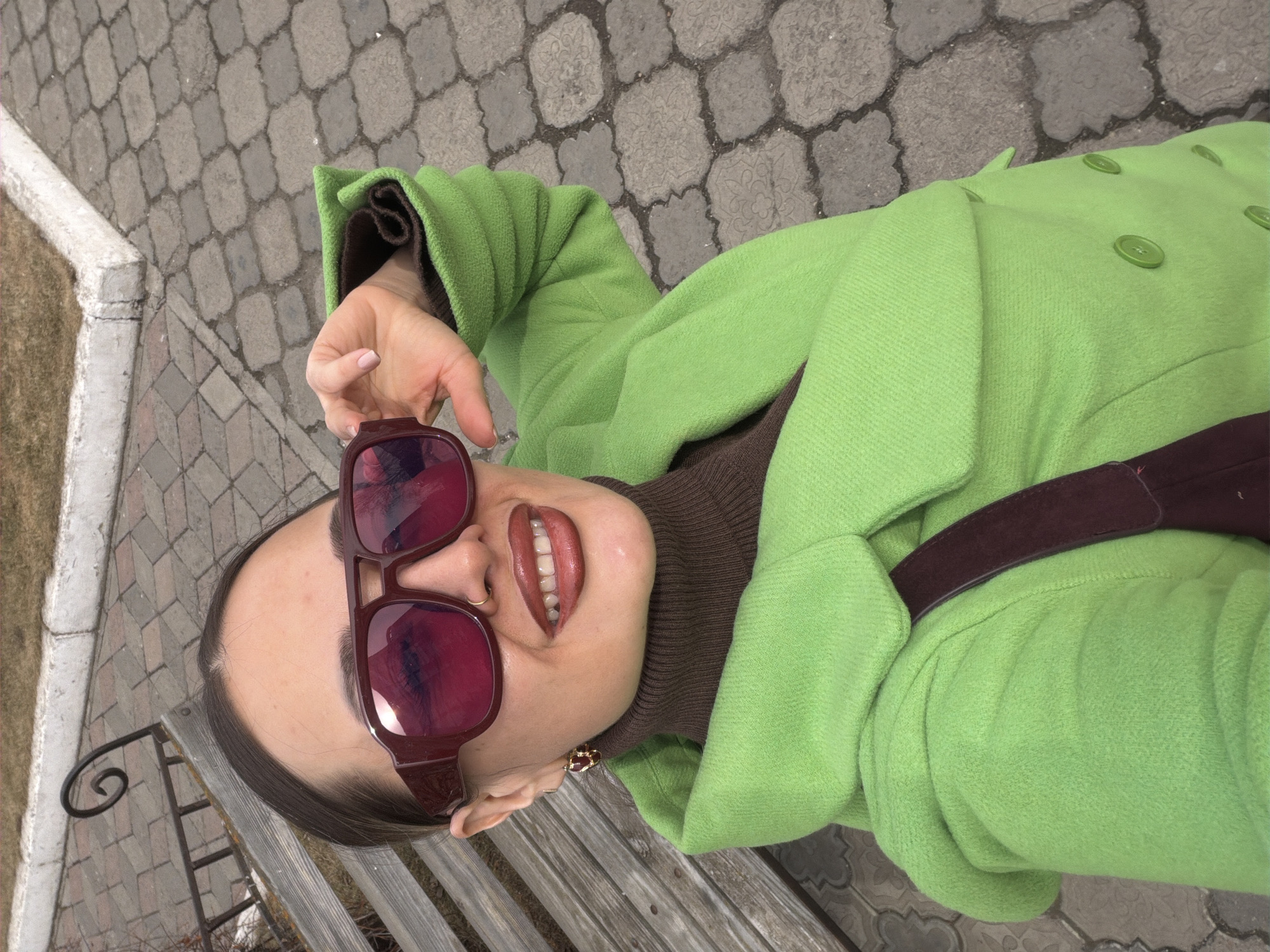 A woman is sitting on a park bench, holding up a pair of sunglasses. She is wearing a brown turtleneck sweater and a green coat. A brown strap is across her chest. The photo appears to be taken in Krasnoyarsk, Russia based on the brick patterned sidewalk.