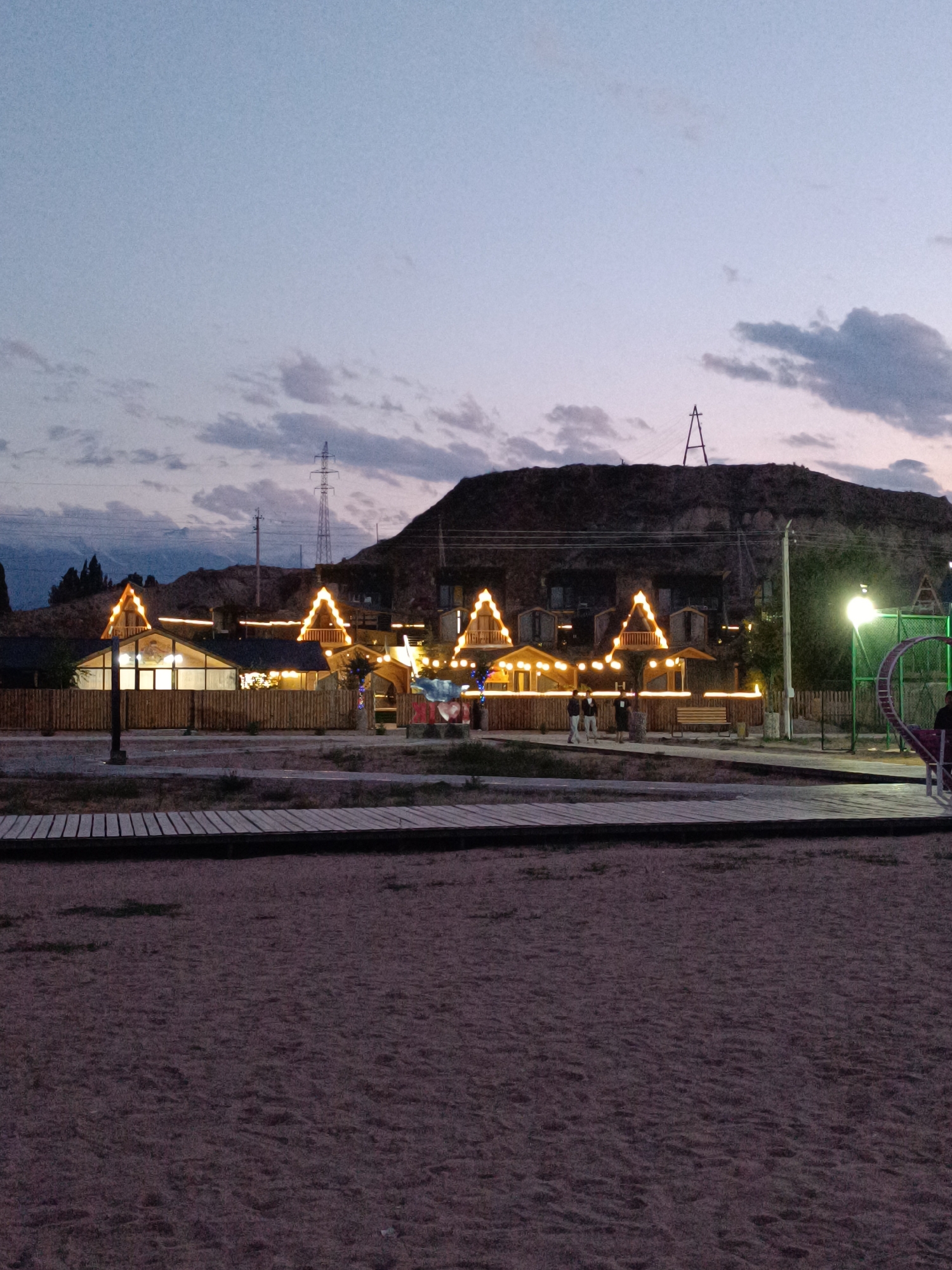 In the city of Tong, Kyrgyzstan, this image captures a twilight scene at a resort or entertainment complex nestled against a hillside. The foreground is dominated by a sandy expanse, with a wooden boardwalk running horizontally across it. Behind the boardwalk, a series of small, chalet-style structures with pointed, illuminated roofs are visible, fronted by a wooden fence. These structures appear to be part of the resort's accommodation or dining facilities, decorated with string lights. In the background, the hillside is rugged and dark, with a tall transmission tower visible on its crest and another, A-frame structure suggesting some form of industrial or historical artifact. A bright, overhead light illuminates a section to the right, near a green metal enclosure that appears to be a playground feature, possibly a hamster wheel for humans, with one person partially visible on it. Several individuals are standing near the buildings, and a wooden bench is placed outside. The sky is a dusky blue, with scattered clouds suggesting the transition from day to night.