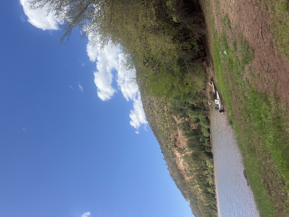 The image depicts a daytime outdoor scene at Ait Ouaarda, Morocco, featuring a body of water adjacent to a vegetated hillside. A clear blue sky with scattered white cumulus clouds dominates the upper portion. A large, still body of brownish-grey water occupies the left side, extending to the lower-left corner. The immediate foreground consists of a dirt bank with patches of green grass and scattered litter, including a white plastic bag. A small, light-colored open boat with two outboard motors is partially beached on the muddy shore near the right-center, with a white object visible on or near it. To the right, a dense cluster of trees with light green foliage lines the bank, casting some shade. On the left, a steep hillside rises from the water, displaying exposed reddish-brown earth interspersed with green shrubs and coniferous trees, some appearing sparse or dry. No individuals are visible.