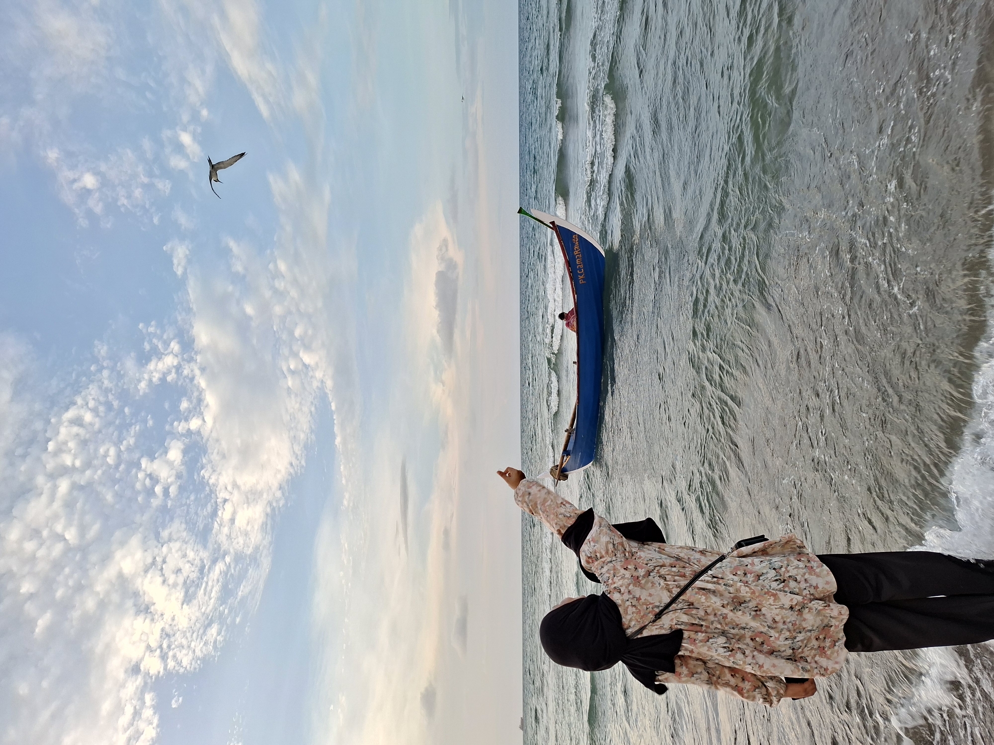 The image shows a beach scene in Lhokseumawe, Indonesia. A woman wearing a black hijab, a floral long-sleeved shirt, and black pants stands on the shore with her arm extended toward a bird flying in the partly cloudy sky. Beside her, a blue and white boat rests partly in the shallow water. The ocean stretches to the horizon.
