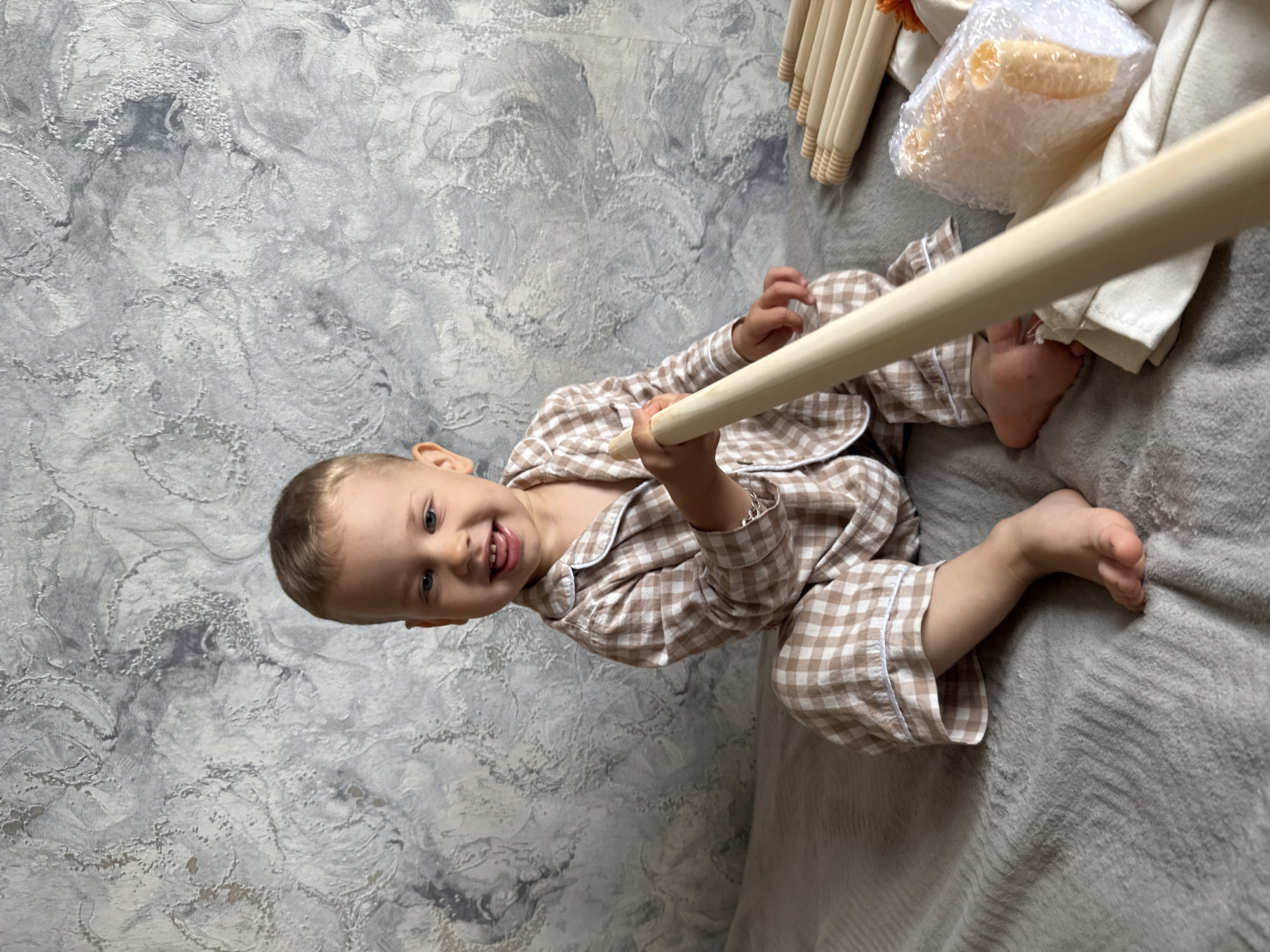 A young child, likely between 2 and 4 years old, sits on a gray textured blanket, perhaps on a bed. The child is wearing a brown and white checkered pajama set. He is holding a wooden rod, partially obscuring the view. To the right of the child, out of focus, there are several other wooden rods, a cushion wrapped in bubble wrap, and a white cloth. The wall is textured with a gray and white marbled design. The scene is likely indoors. There is no information in the image that would indicate it is in Rusanovo, Russia.