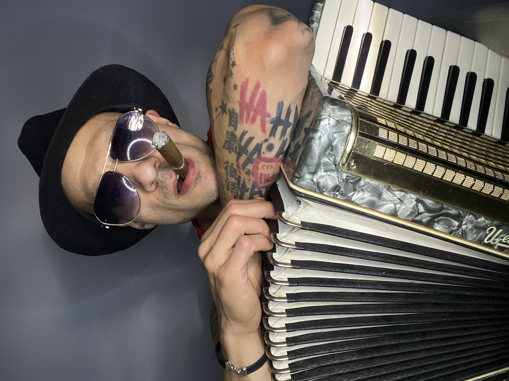 A man is angled on his side in the photograph, wearing a black fedora and sunglasses, and smoking a cigar. His right arm, which has multiple tattoos, is bent and raised. He is holding the side of an accordion with his right hand. The accordion has black and white keys. The backdrop is a solid gray color. The photo was not taken in Pleven, Bulgaria.