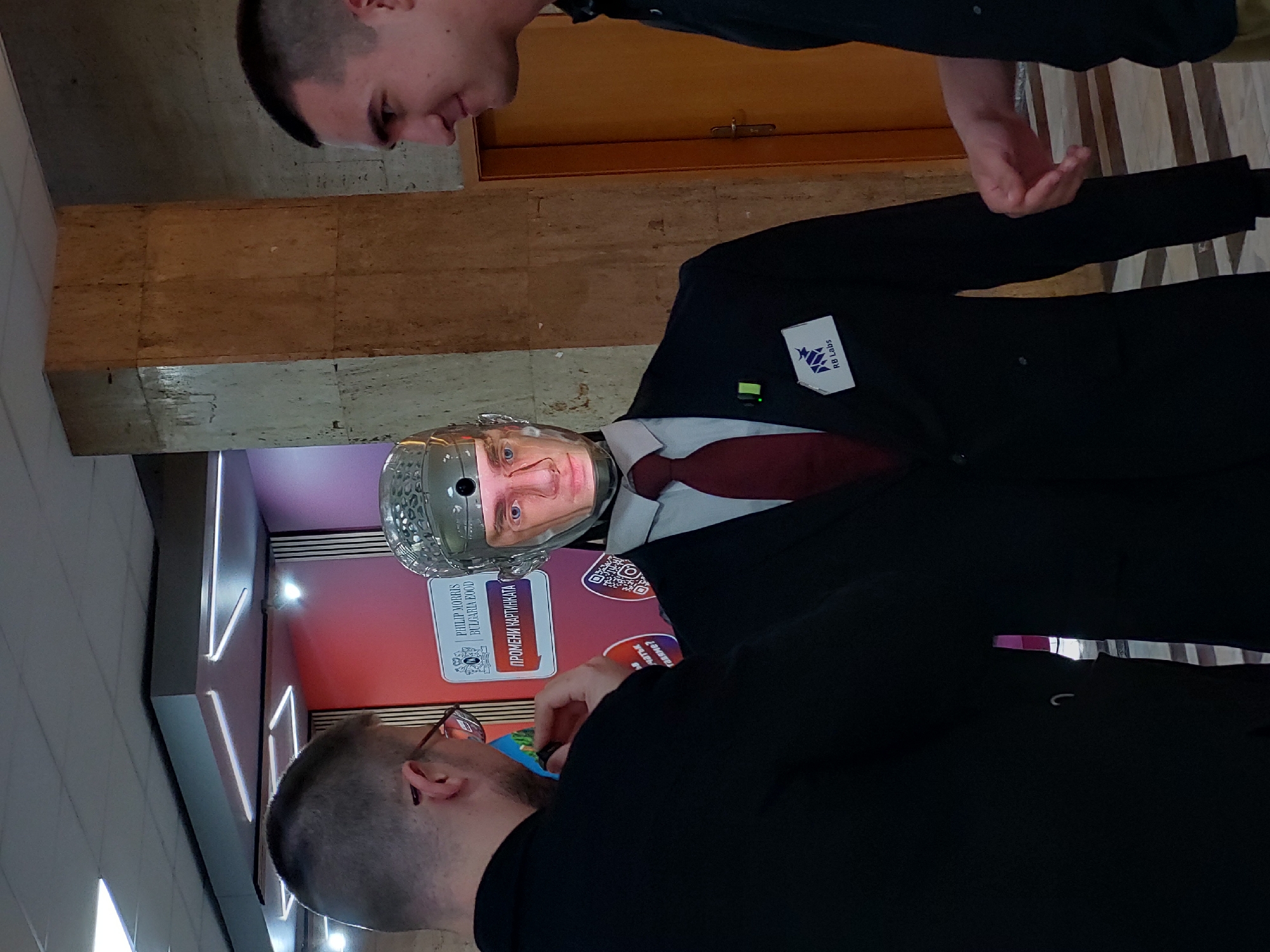 The image displays an indoor scene featuring two men interacting with a humanoid robot, likely at an exhibition or public event.
The central subject is a robot dressed formally in a black suit, a white shirt, and a dark red tie. Its head is unique, consisting of a transparent, dome-shaped casing that encloses a screen displaying a man's face, notably resembling Vladimir Putin. A white rectangular badge with the text "RB Labs" and a blue logo is pinned to the robot's left lapel, alongside a small green indicator light.
Two men are observing the robot. In the upper part of the frame, a man with short, dark hair is seen looking down at the robot, with his right hand extended towards it. In the lower foreground, another man, wearing glasses and with short hair, is shown from behind, also intently looking at the robot.
The background consists of a wall clad in light brown, rectangular tiles, and a wooden door. To the left of the robot, a brightly lit display stand is visible. This stand features a red and purple gradient background, multiple QR codes, and text primarily in Cyrillic. Visible text includes "ПРОМЕНИ КАРТИНАТА" (Change the picture/painting) and a logo with "PHILIP MORRIS BULGARIA О.O.D." (or similar corporate designation). The overall setting suggests a technological demonstration or an educational display in a public or corporate environment.