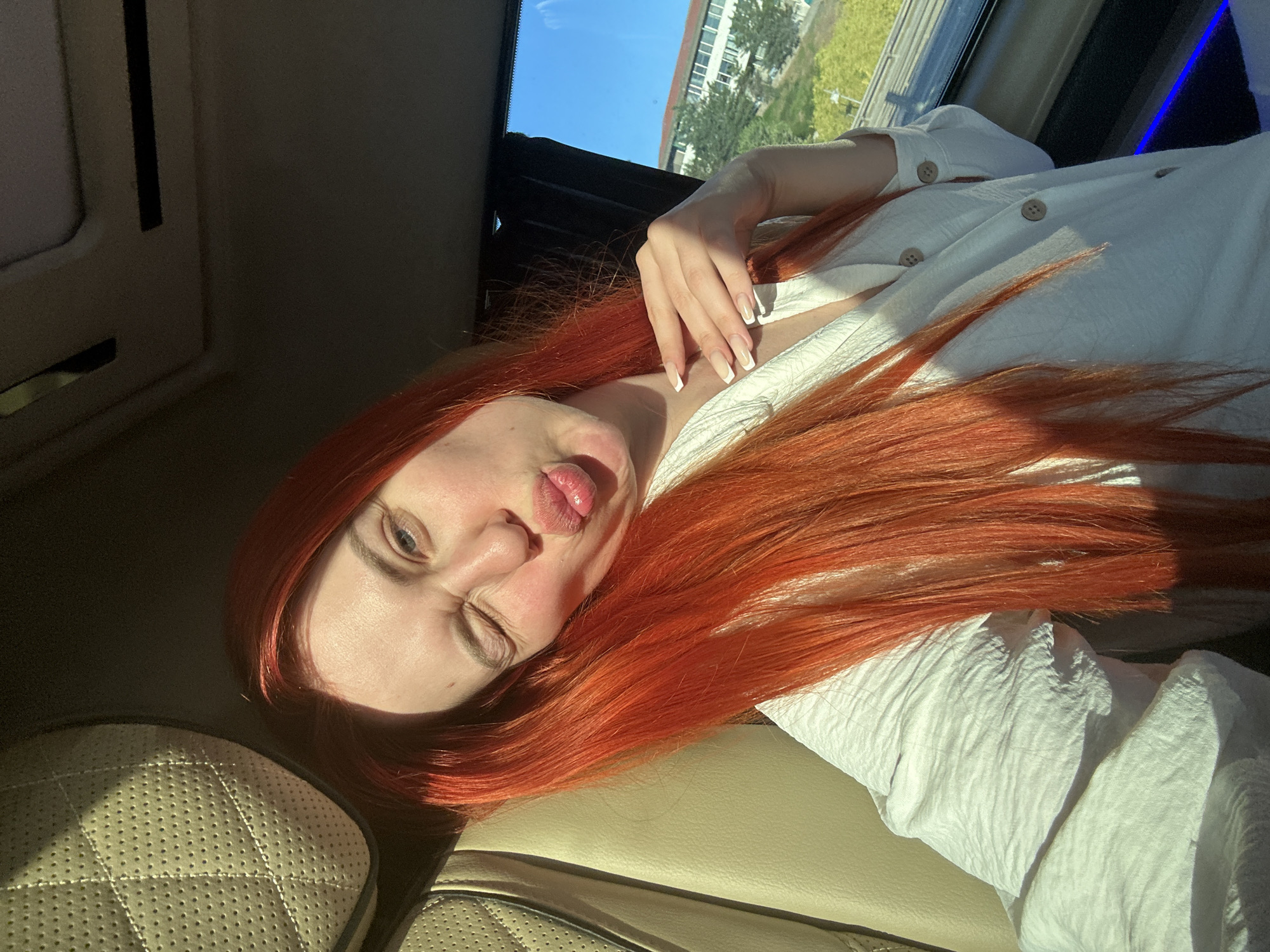 A woman with long, bright orange hair is taking a selfie inside a vehicle. She is winking and puckering her lips. She is wearing a white shirt with buttons. The interior of the vehicle appears to be luxurious, with quilted cream-colored upholstery visible on the left. Sunlight is streaming in from the right, casting shadows. Outside the window, a building and trees are visible, suggesting an urban or suburban setting. The exact location in Eyüpsultan, Turkey cannot be determined from the image alone.