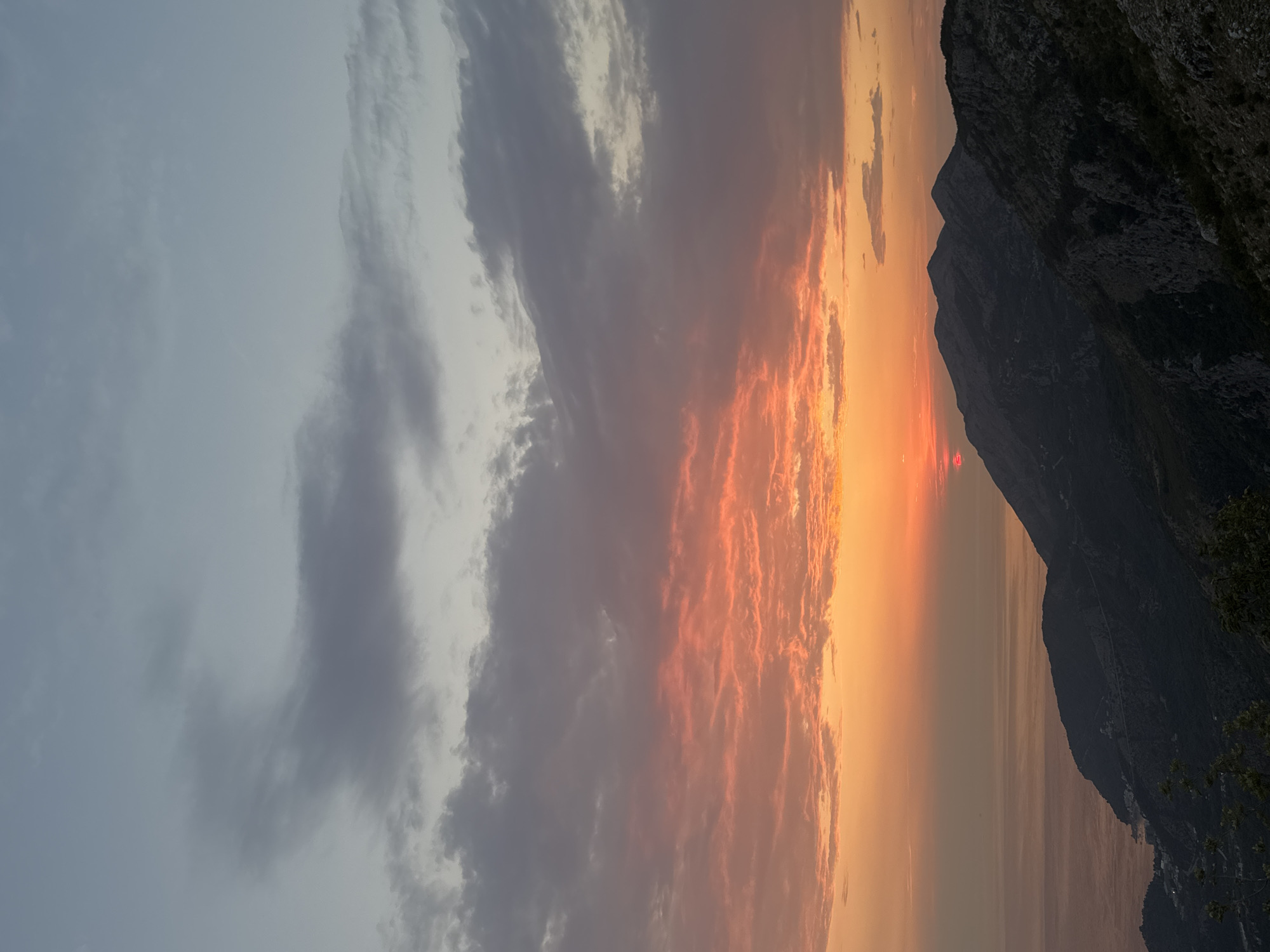 This is an outdoor, landscape photograph taken at dusk in Gluhi Do, Montenegro. The image captures a dramatic sunset over a mountainous coastal region. The sky is filled with a gradient of colors, transitioning from a pale blue at the top to vibrant oranges and reds near the horizon, illuminated by the setting sun, which is partially visible just above a mountain peak. Fluffy clouds are scattered across the sky, some tinged with pink and orange from the sunset's light, while others are a darker grey.
In the foreground, rugged, dark mountains dominate the lower portion of the image. Their slopes are covered in dense vegetation, with visible rock formations and steep inclines. The terrain suggests a challenging and wild landscape. To the left, the sea is visible, its surface reflecting the soft colors of the sky. Along the coastline, faint lights indicate a small settlement or village nestled between the mountains and the water. The overall impression is one of natural beauty and serene twilight. No people or animals are visible in the scene.