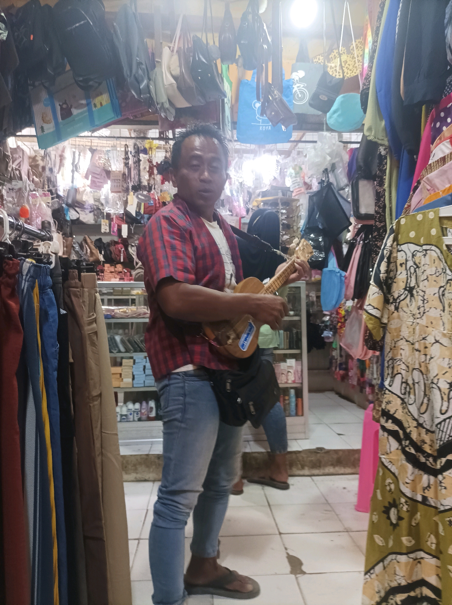 A man stands in a market stall in Sumuragung, Indonesia, playing a ukulele. He is wearing a red and blue plaid shirt, blue jeans, and dark sandals. A black bag is slung across his chest. The stall is filled with merchandise, including clothing racks, shelves of products, and various bags hanging from the ceiling. The floor is tiled. The lighting is bright, suggesting an indoor setting with overhead illumination. The background shows more stalls and a glimpse of the market's interior.