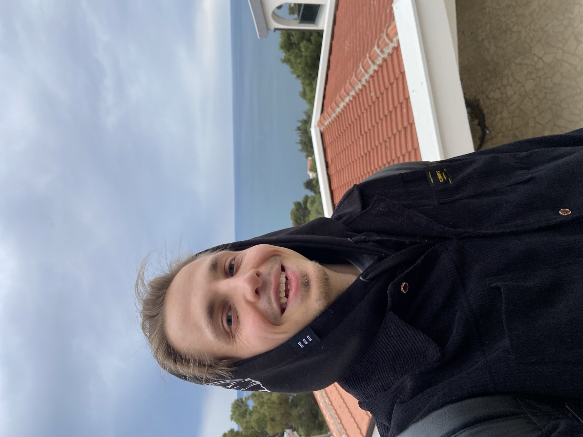 A selfie photograph shows a man with light hair and a slight beard, wearing a black hooded jacket, smiling towards the camera. He is standing on a red-tiled roof near a white building structure, likely in Šušanj, Montenegro, as a sea and a cloudy sky are visible in the background. Vegetation can be seen in the distance.