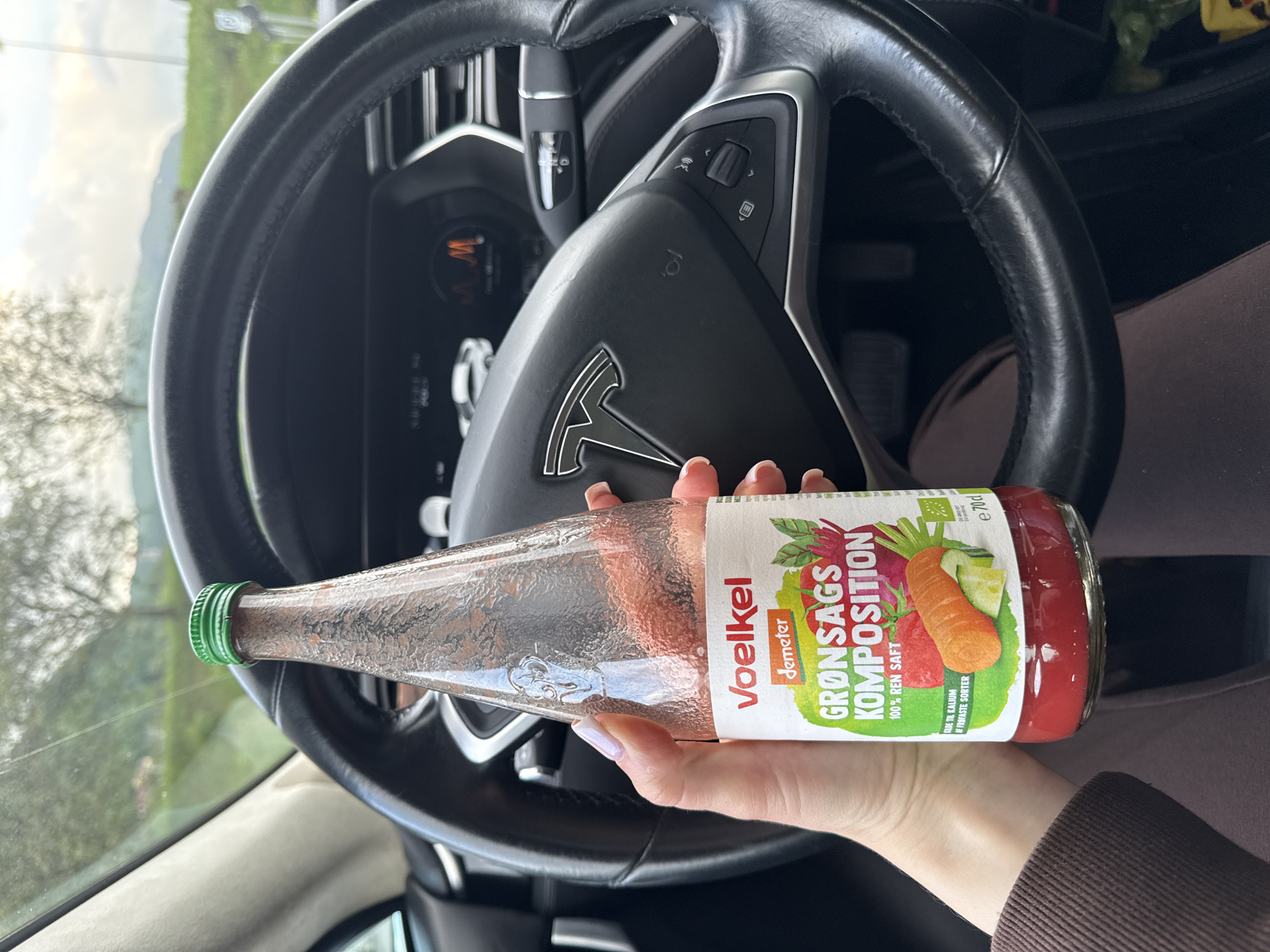 A woman's hand, with pale skin and manicured nails, holds a glass bottle of Voelkel Grønsags Komposition juice. The bottle has a green cap and a white label with red and green text and illustrations of vegetables. The juice inside is a deep red color. The bottle is held in front of a black steering wheel of a Tesla car, with the car's dashboard visible in the background. The scene is indoors, likely inside a car, with a blurred outdoor background of trees and a landscape. The lighting suggests it is daytime.