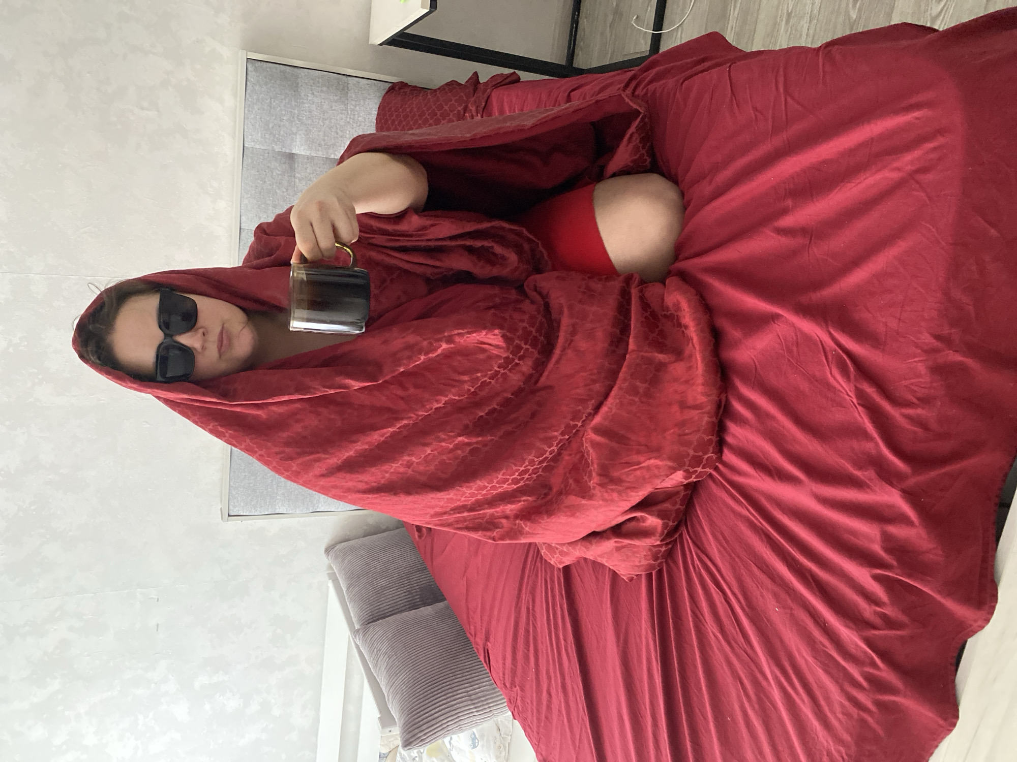 A woman lies in bed, covered in a red blanket, with only her face visible through large black sunglasses. She is holding a dark-colored mug in her right hand. The bed has red sheets and is located in a room with light-colored walls and a gray headboard. A nightstand is visible next to the bed. A red sock is partially visible.