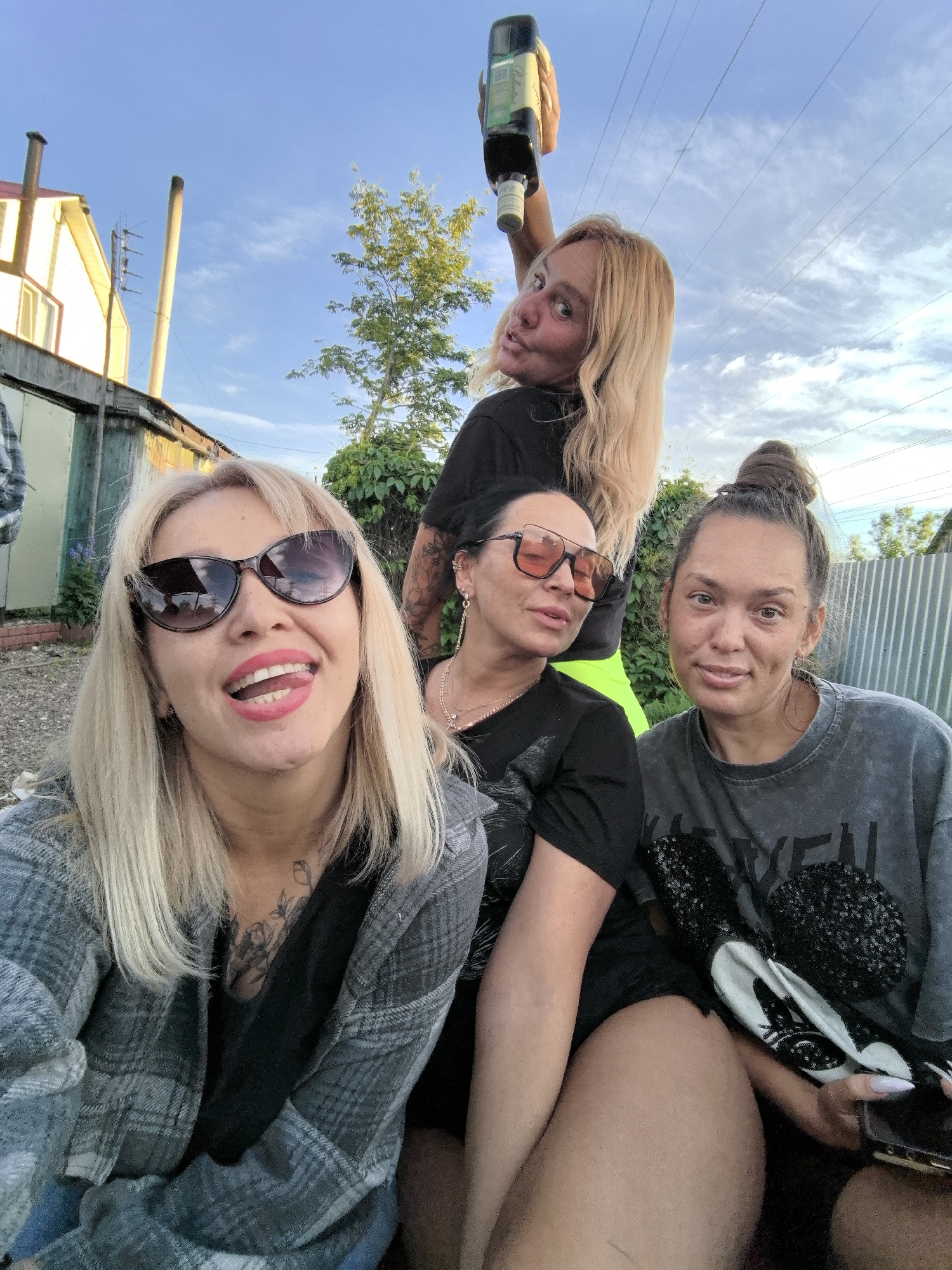 Four adult women are captured in a casual outdoor photo, likely a selfie, in Noviy, Russia.
The woman on the far left has blonde hair, wears sunglasses, a black top, and a plaid shirt, and smiles playfully with her tongue slightly out. A tattoo is visible on her chest. Next to her, slightly behind, another woman with dark hair wears orange-tinted sunglasses and a black top. A tattoo is visible on her right arm.
At the top of the frame, a third woman with blonde hair, dressed in a black top and neon green shorts, holds a dark green Jägermeister bottle aloft with one hand, looking over her shoulder towards the camera with a playful expression. The bottle clearly shows "Jägermeister" and "HERBAL LIQUEUR" on its label.
On the right side of the image, the fourth woman has dark hair styled in a bun and wears a grey t-shirt with sequins and faded white text. She holds a cell phone and has a Mickey Mouse-themed sequined bag resting beside her. Her fingernails are long and white.
Parts of a fifth person, including an arm and plaid clothing, are visible on the far left.
The setting appears to be a residential backyard or a casual outdoor gathering space. In the background, there's a light yellow house with two tall, slender chimneys or pipes and an antenna. Other outbuildings with corrugated metal siding are also visible, along with green trees, some power lines in the clear blue sky, and a metal fence. The sky is bright with scattered clouds, indicating daytime, possibly late afternoon or early evening. The overall mood is relaxed and informal.