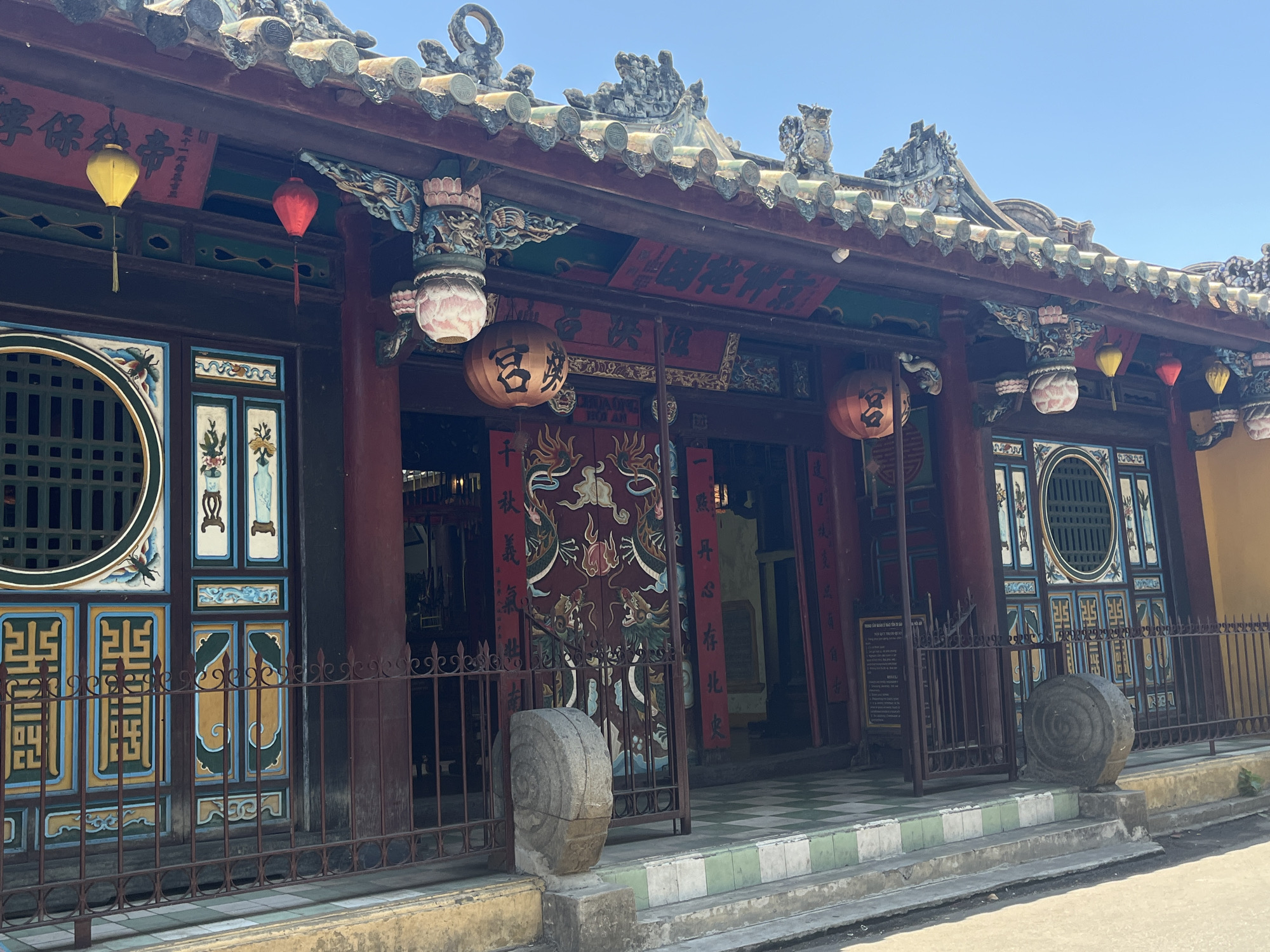 This media file captures the ornate facade of a traditional building, likely a temple or communal house, set against a clear blue sky in Hội An, Vietnam. The building features a dark wooden structure with a multi-tiered, tiled roof adorned with intricate ceramic figures and decorative carvings, including mythical creatures.
The facade is richly decorated with painted panels in blue, green, and gold, showcasing geometric patterns, floral motifs, and what appears to be a spider or scorpion on one of the window sections. Numerous colorful lanterns, including yellow, red, and large orange ones bearing Chinese characters such as "宮" (palace/temple) and "殿" (hall/palace), hang from the eaves.
The main entrance features prominent red double doors with painted dragon designs. Above these doors, a large red horizontal banner displays Chinese characters, which, along with other text, include "享保錢帝" (possibly relating to imperial coinage or a historical era) and "HOI AN" on a smaller sign below. Vertical red couplets flanking the main doors read "千秋義" (eternal righteousness) and "心存北史" (heart remembers Northern history).
In front of the building, a low, dark metal fence encloses a small area, and two large, round stone carvings with spiral patterns flank the entrance steps. The steps themselves are tiled with a distinctive green and white checkerboard pattern. The bright, clear daylight suggests it is midday or early afternoon, highlighting the vibrant colors and detailed craftsmanship of the structure. No people are visible, emphasizing the architectural beauty of the site.