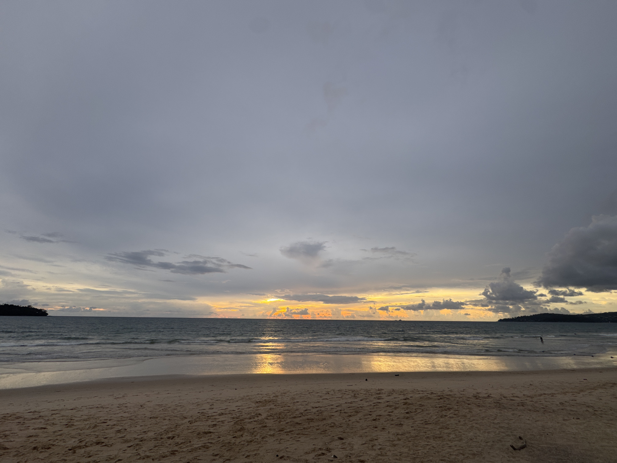 This is a wide-angle, eye-level, outdoor photograph of a beach at dusk. The sky is filled with a mix of dark and light gray clouds, with a bright, colorful sunset peeking through the clouds near the horizon. The ocean is a deep blue-gray with gentle waves lapping at the shore. The sand on the beach is a light tan color, with some scattered debris. In the distance, there are two small islands, one on the left and one on the right, both covered in dark green vegetation. On the right side of the image, a single person can be seen wading in the shallow water. The image captures a serene and natural landscape, indicative of a coastal area in Choeng Thale, Thailand, likely during the twilight hours.