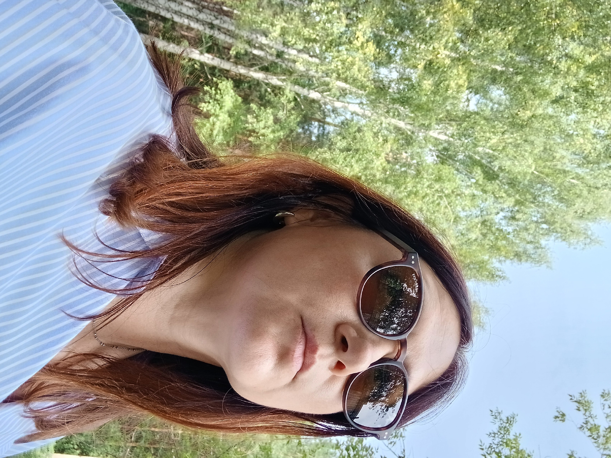 A woman wearing sunglasses and a light blue and white striped button-down shirt is pictured from the shoulders up, looking upwards and to her right. She has brown hair and is wearing a thin silver chain necklace and small gold hoop earrings. The background is out of focus and shows green trees and a blue sky, suggesting an outdoor setting in a wooded area. There are no visible indicators of the specific city of Osa, Russia.