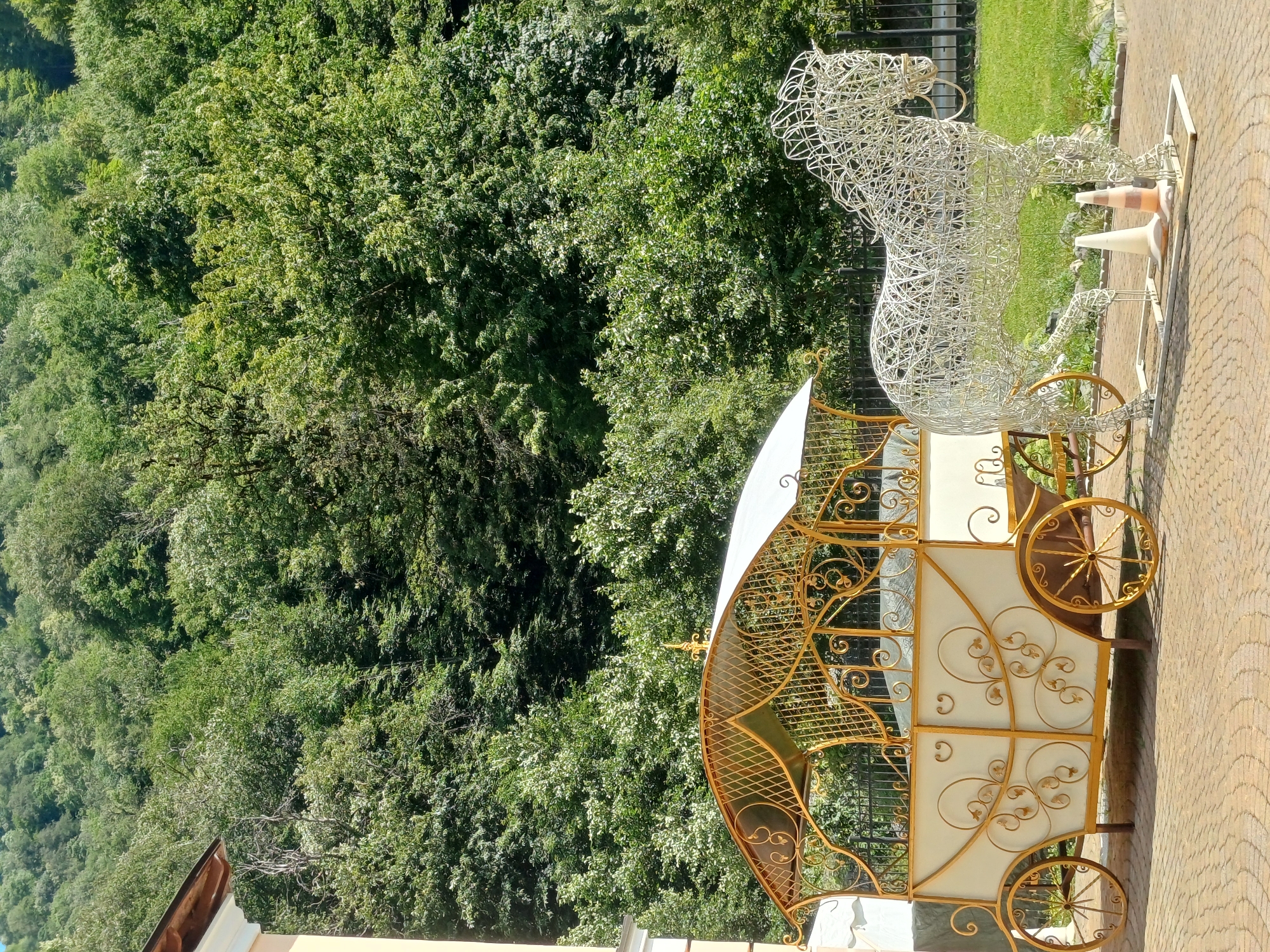 The image depicts an outdoor scene in Estosadok, Russia. In the foreground, a decorative horse sculpture made of white wire stands on a paved pathway. To its left is a carriage, also decorative, with a cream-colored body and ornate gold scrollwork. The carriage has a white canopy and gold wheels. The pathway is made of interlocking brick pavers. Behind the carriage and horse, a lush, dense green forest covers a hillside. The trees are vibrant and full, indicating a summer or late spring setting. The area appears to be a park or garden, possibly near a building, as evidenced by a sliver of a light-colored building with a wooden roofline visible in the upper left corner. The lighting suggests it is a sunny day.