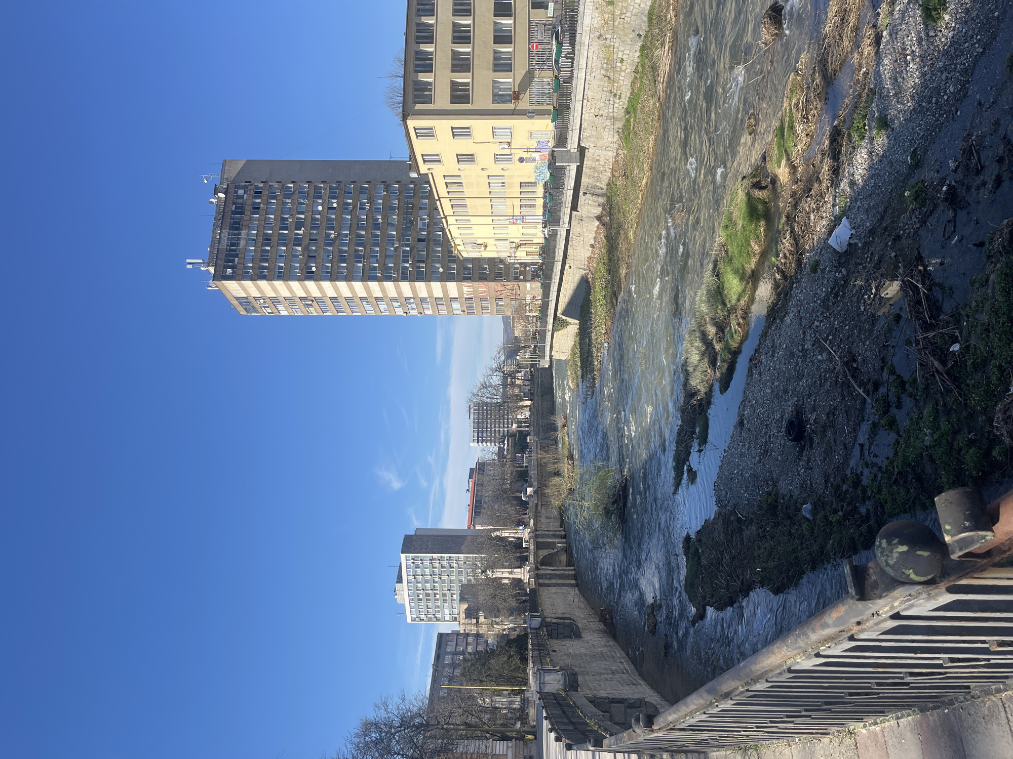 The image depicts a river flowing through an urban environment, possibly in Gabrovo, Bulgaria, characterized by a mix of architectural styles and infrastructural elements. The river, bordered by concrete and stone embankments, exhibits varying water levels and flow. River banks contain gravel, moss and litter. On the left side of the image, several buildings are visible. A tall, gray high-rise building with numerous windows dominates the skyline, followed by a beige-colored building with a similar grid pattern of windows. Several buildings of varying sizes can be seen in the distance. The sky is clear and blue. In the foreground, a metal railing or fence is present, providing a boundary between the viewer's perspective and the river below.