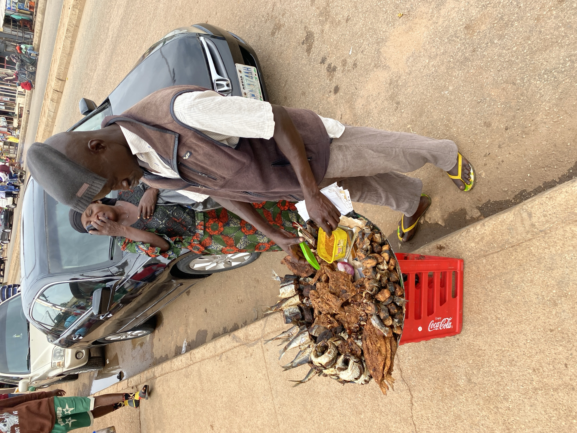 A street vendor in Bukuru, Nigeria, serves a female customer at a black car. The vendor, wearing a gray beanie, brown vest, and gray pants, offers cooked fish displayed on a large platter set on a red Coca-Cola crate. The woman, in a colorful patterned dress and head wrap, appears to be on her cell phone while making her order. The license plate on the car reads "ABUJA FL-174AH". A second car is parked next to the black car. Other people and structures are visible in the background. The setting is on a paved street during daylight.