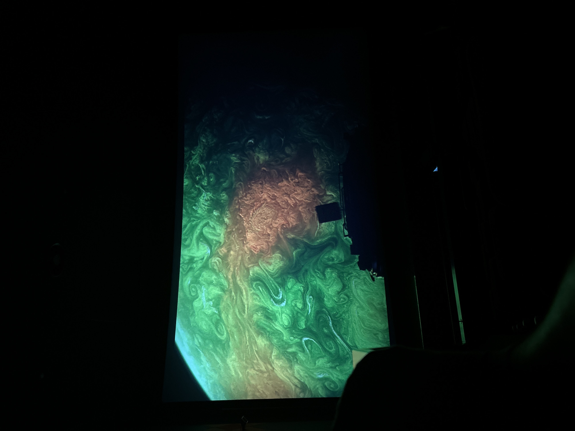 The image shows a planetarium screen displaying a swirling, colorful image resembling a planet. The predominant colors are green, orange, and brown, suggesting a swirling gas giant. The surrounding environment is dark, indicating a darkened planetarium theater. The image appears to be taken from a seated position, with hints of seats and potentially a person's arm visible in the foreground.
Although the prompt includes Burgas, Bulgaria, there is nothing in the image that confirms this location. Therefore, Burgas is not part of the forensic description.