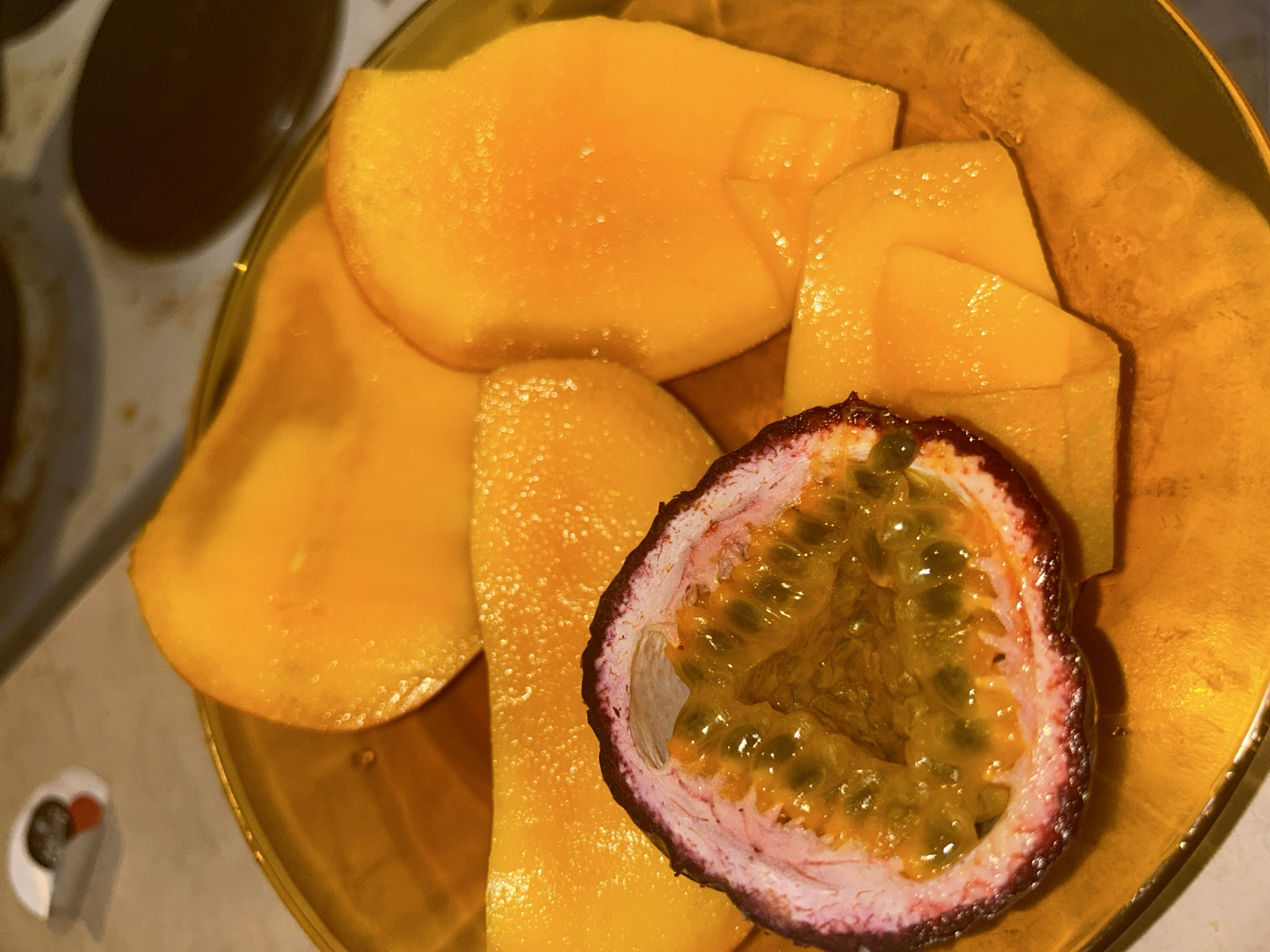 A top-down shot shows a gold-colored plate containing slices of mango and half of a passion fruit. The mango slices are bright yellow-orange and vary in size and shape. The passion fruit half has a dark purple and pink rind, revealing a yellowish-brown pulp filled with seeds. The plate sits on a surface, partially revealing a counter or tabletop on the left side of the frame. A glimpse of what appears to be a stovetop can be seen in the upper left corner.
It is not possible to determine the location in Varna, Bulgaria, or any information about people, actions, interactions, or the environment beyond what is explicitly shown in the image.