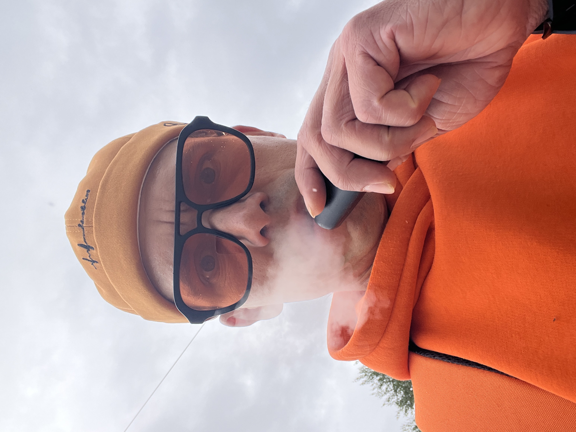 An overhead, slightly angled shot shows a person exhaling vapor from a vaping device against an overcast sky in Chaikovsky, Russia. The image is oriented with the person's head on the left and their body extending to the right.
The central subject is an individual, likely male, wearing an orange beanie with dark, illegible cursive embroidery on the side. They are also wearing black-framed glasses with amber-tinted lenses, through which their eyes are visible. A black vaping device is held to their mouth by their right hand, and a cloud of white vapor is seen emanating from around their mouth and nose.
The person is dressed in a vibrant orange hoodie or sweatshirt, the textured fabric of which is visible, especially on the right side of the frame and around their neck. The right hand, holding the vape device, has visibly wrinkled skin.
The background is dominated by a light gray, cloudy sky, indicating an overcast day. In the lower right corner of the broader image, a hint of green foliage, possibly from trees or bushes, can be seen, suggesting an outdoor, possibly natural, setting. The overall mood is casual.