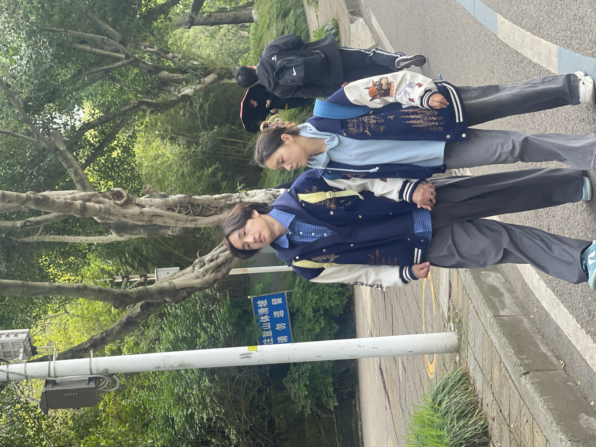 The image shows a city street scene, likely in the Chongqing High-tech Zone, China. Two young individuals, dressed in matching letterman-style jackets, are standing or walking on a sidewalk next to a curb. A signpost with Chinese characters is visible, partially obscured by the person in the foreground. Trees with exposed roots line the street, creating a green canopy overhead. In the background, two other people are walking, one with a backpack. The sidewalk is marked with painted lines, suggesting it's a designated pedestrian path. The overall environment suggests a residential or park-like area within the city.