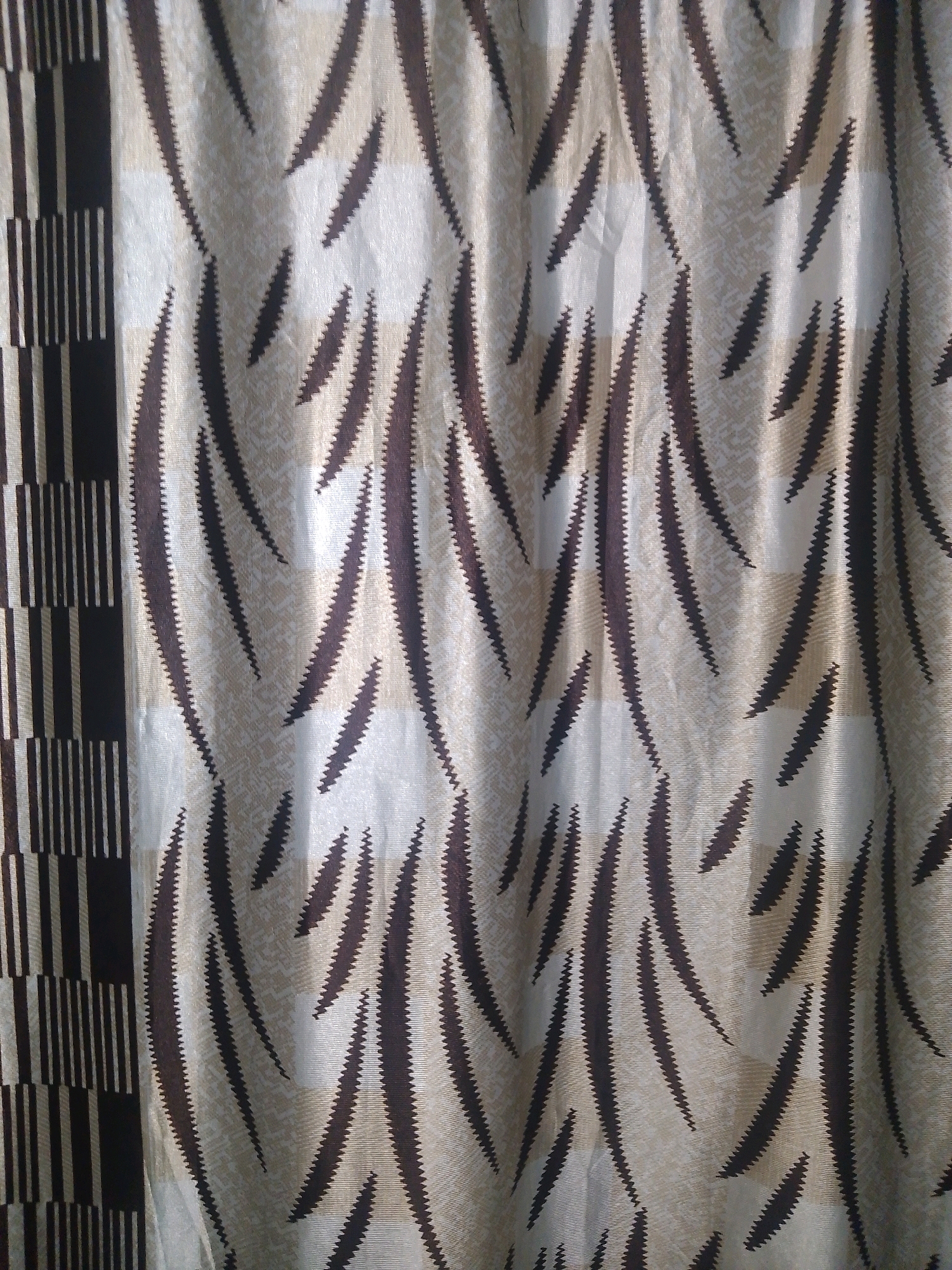 A close-up, eye-level, medium shot captures a curtain with a tiger-stripe pattern. The curtain is made of fabric with a textured, woven surface. The dominant pattern consists of elongated, dark brown, jagged shapes that resemble tiger stripes, set against a lighter, beige background. To the left of the frame, a vertical section of the curtain displays a contrasting geometric pattern of alternating dark brown and beige vertical stripes and squares. The fabric exhibits some folds and wrinkles, indicating it is hanging or draped. No people, animals, or specific location details are visible.
