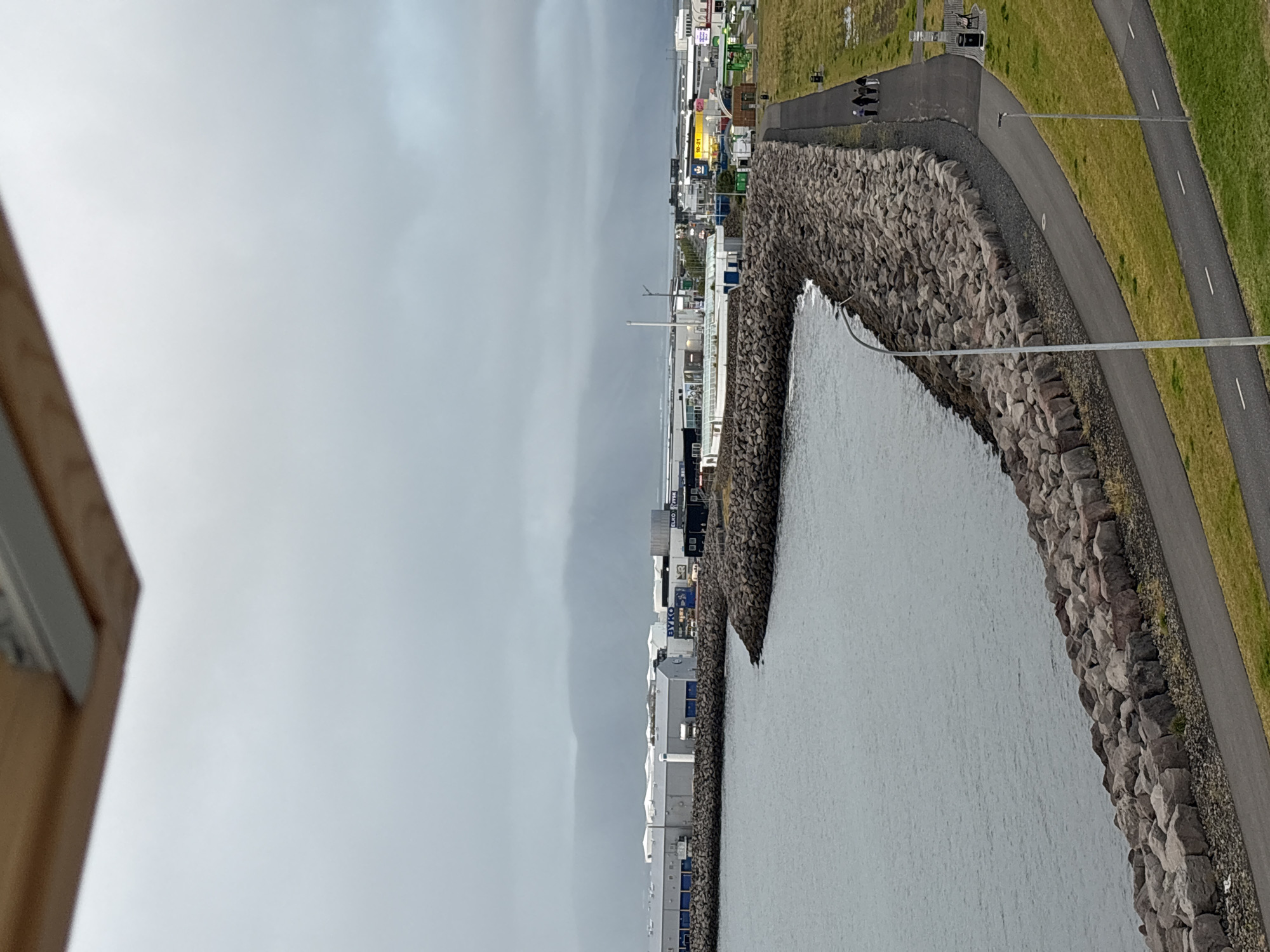 The image captures a cloudy day view of the Reykjavik coastline. In the foreground, a paved walkway follows a curved breakwater constructed from large rocks. To the right of the walkway, a strip of green grass is visible, punctuated by two streetlights. A group of four individuals are walking along the walkway. Further into the scene, industrial buildings and commercial establishments are visible along the coastline, with some structures displaying signage. In the distance, a hazy mountain range forms the backdrop under the overcast sky. The water is a calm, muted gray. The overall scene suggests an urban coastal environment with recreational pathways and industrial infrastructure.